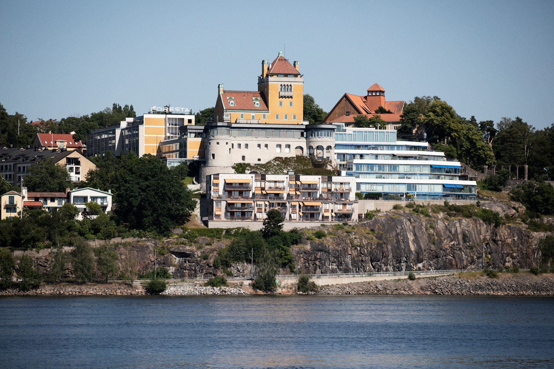 Ett gult slott med torn ligger på en klippa ovanför en blå flod, omgivet av gröna träd och moderna byggnader.
