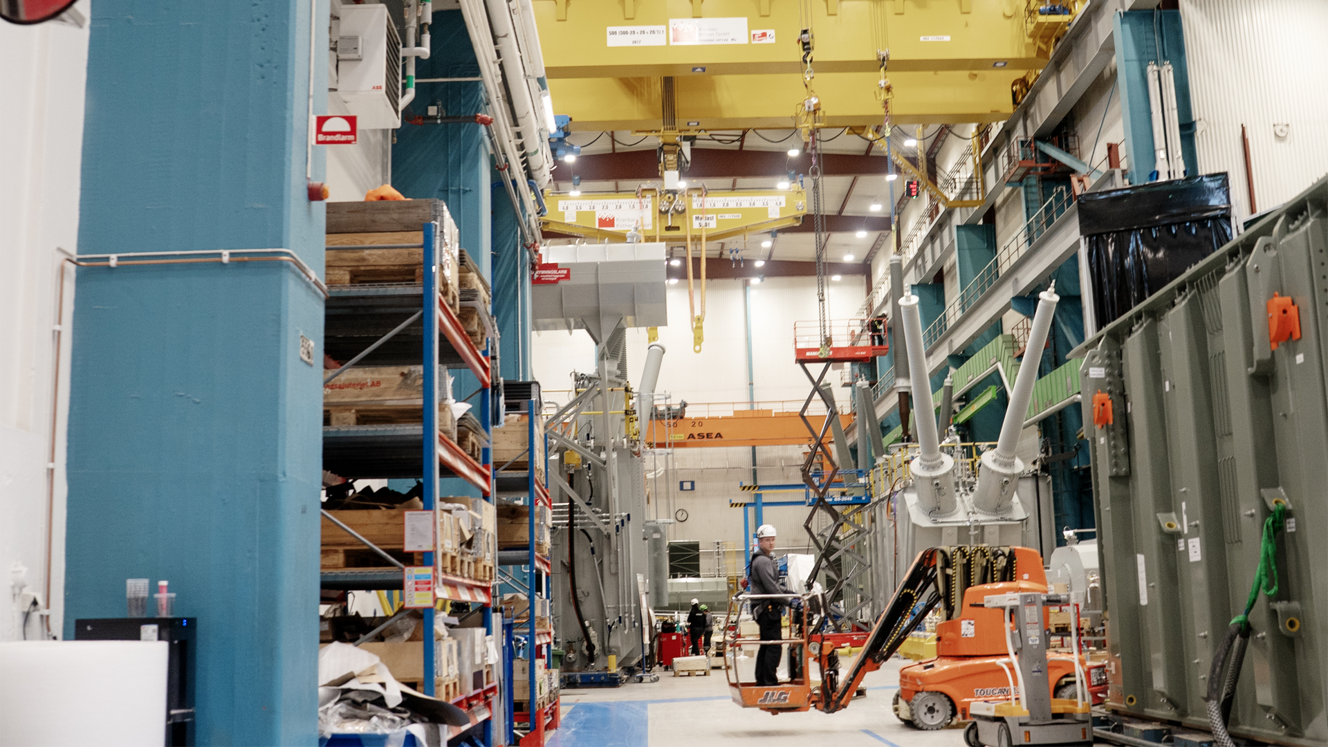 Industriell fabrikshall med gul lyftkran, blå och gröna väggpaneler, orange arbetsutrustning och arbetare som utför monteringsarbete på en stor elektrisk transformator eller kraftutrustning.