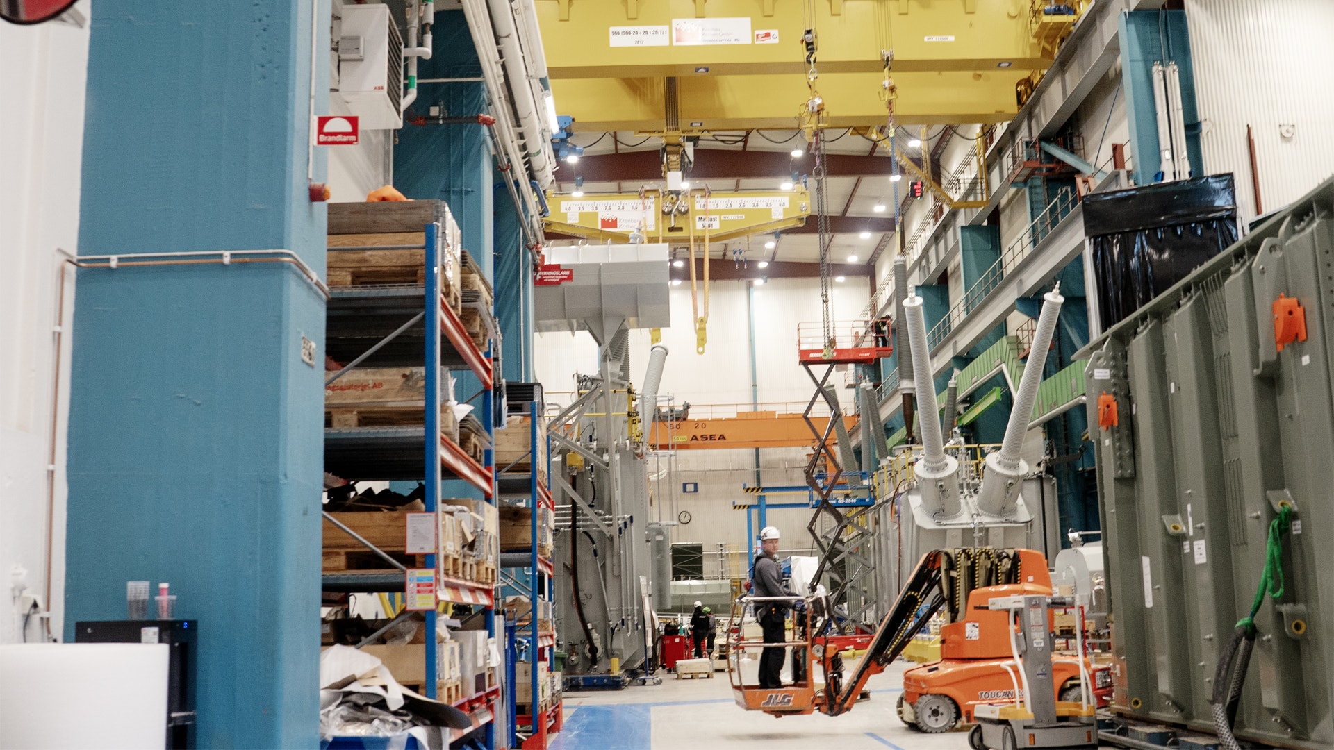 Industriell fabrikshall med gul lyftkran, blå och gröna väggpaneler, orange arbetsutrustning och arbetare som utför monteringsarbete på en stor elektrisk transformator eller kraftutrustning.
