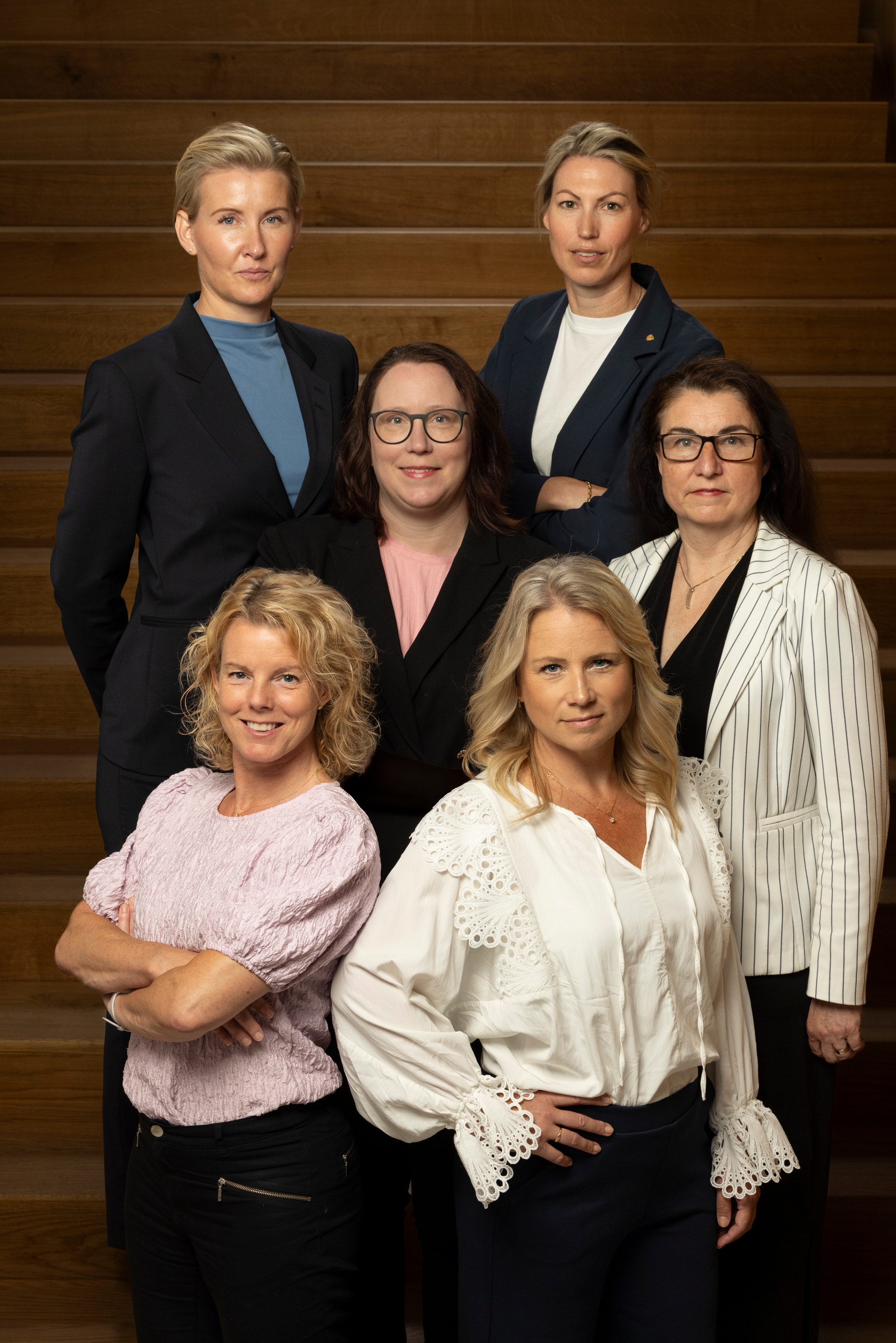Gruppfoto av sex kvinnor i professionell miljö mot en trävägg. De är klädda i affärsklädning och poserar tillsammans för kameran.