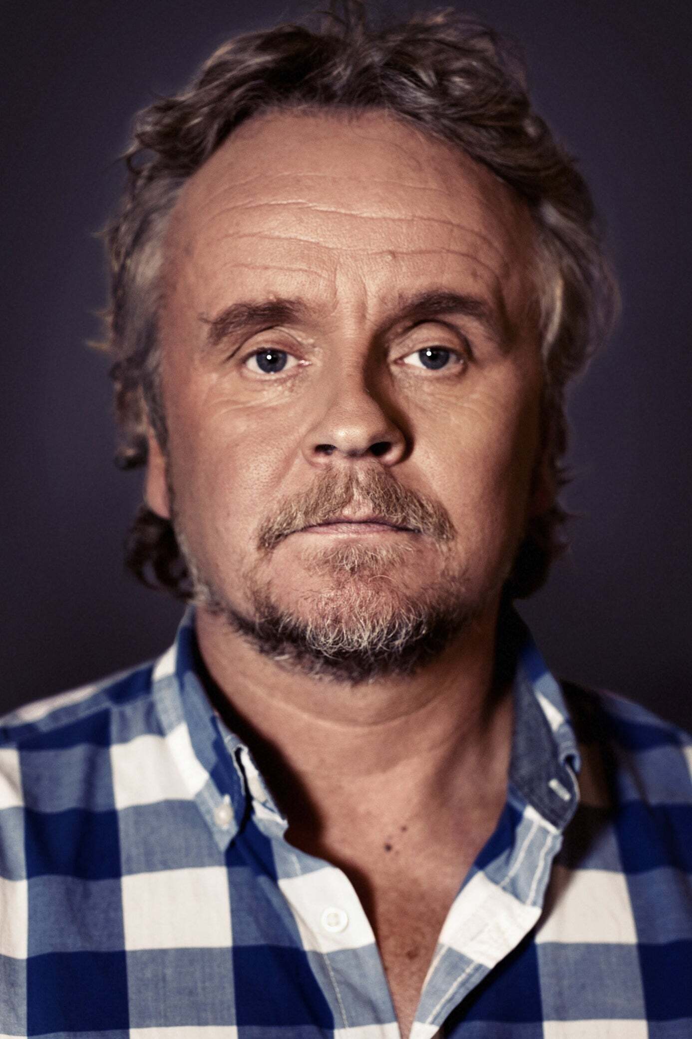 Porträtt av en man med mörkt hår och mustasch, klädd i en blå och vit rutig skjorta mot en mörk bakgrund.