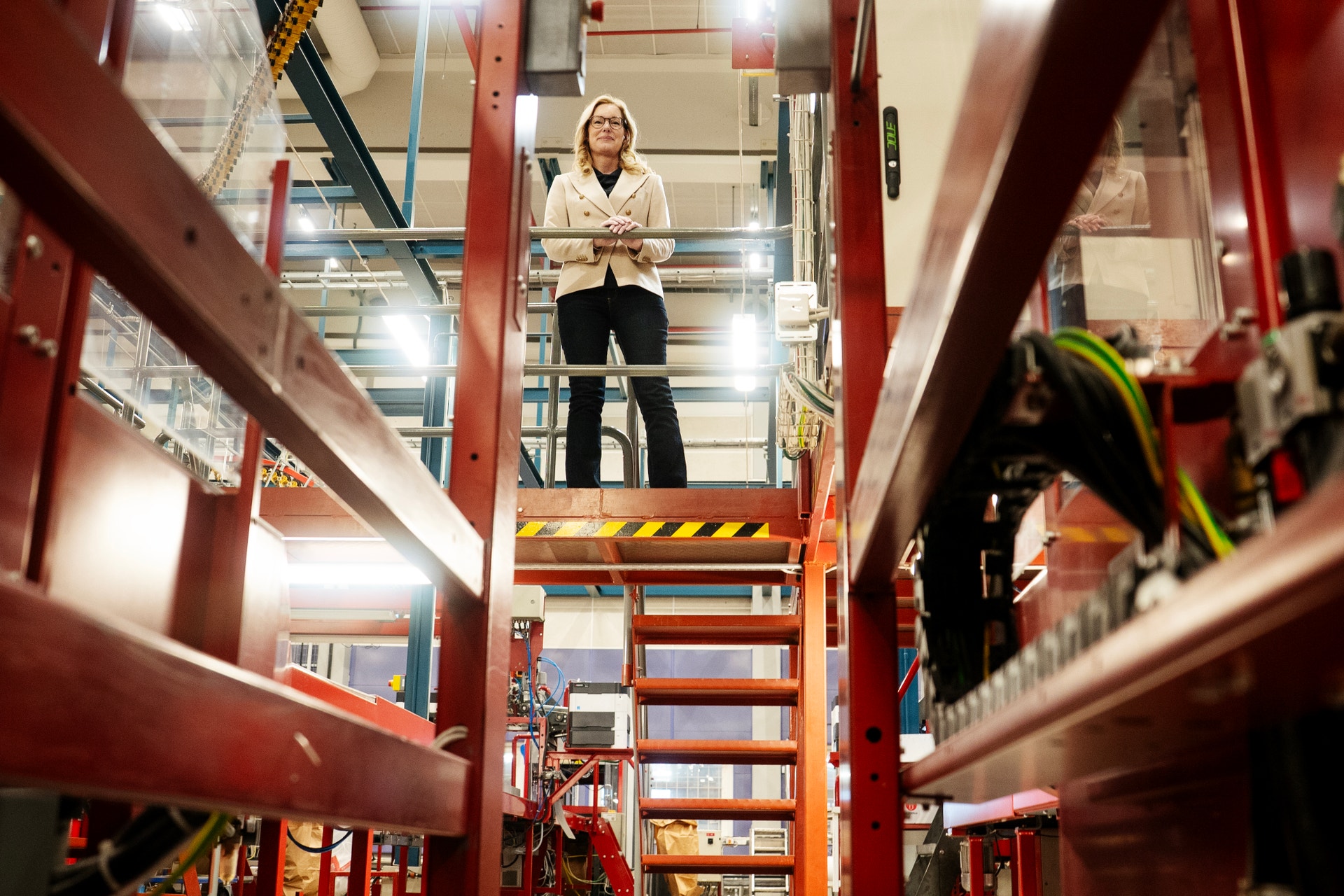 Kvinna står på en upphöjd industriplattform med röda säkerhetsgrindar i ett lager eller industriell miljö.
