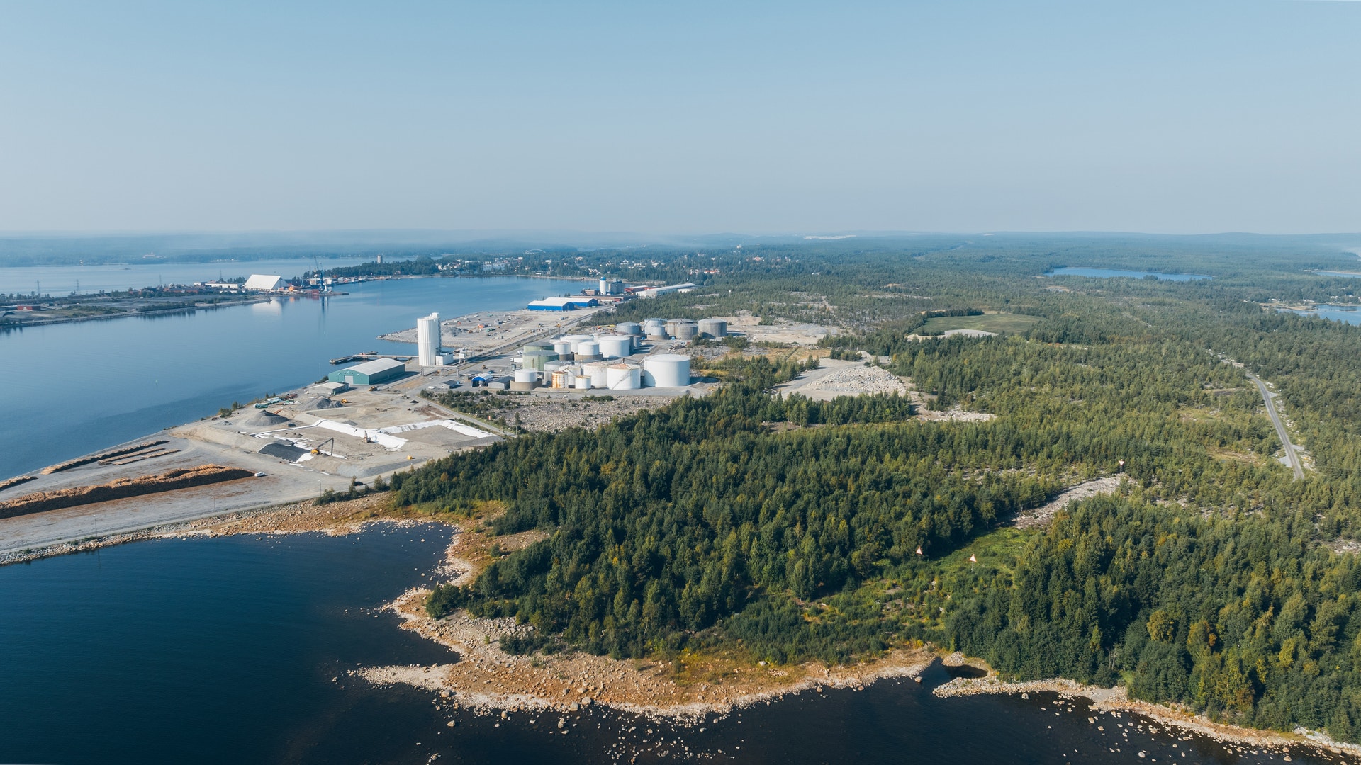 Flygfoto över en industrianläggning med vita tankar och byggnader vid en blå vattenbody, omgiven av grön skog.