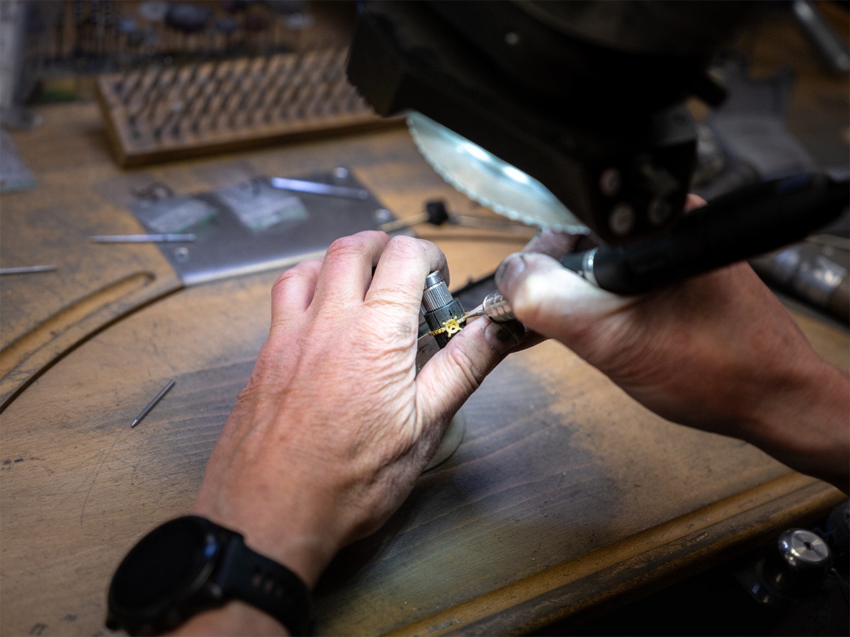 En guldsmeds händer arbetar med en guldring på en arbetsbänk omgiven av verktyg och material.
