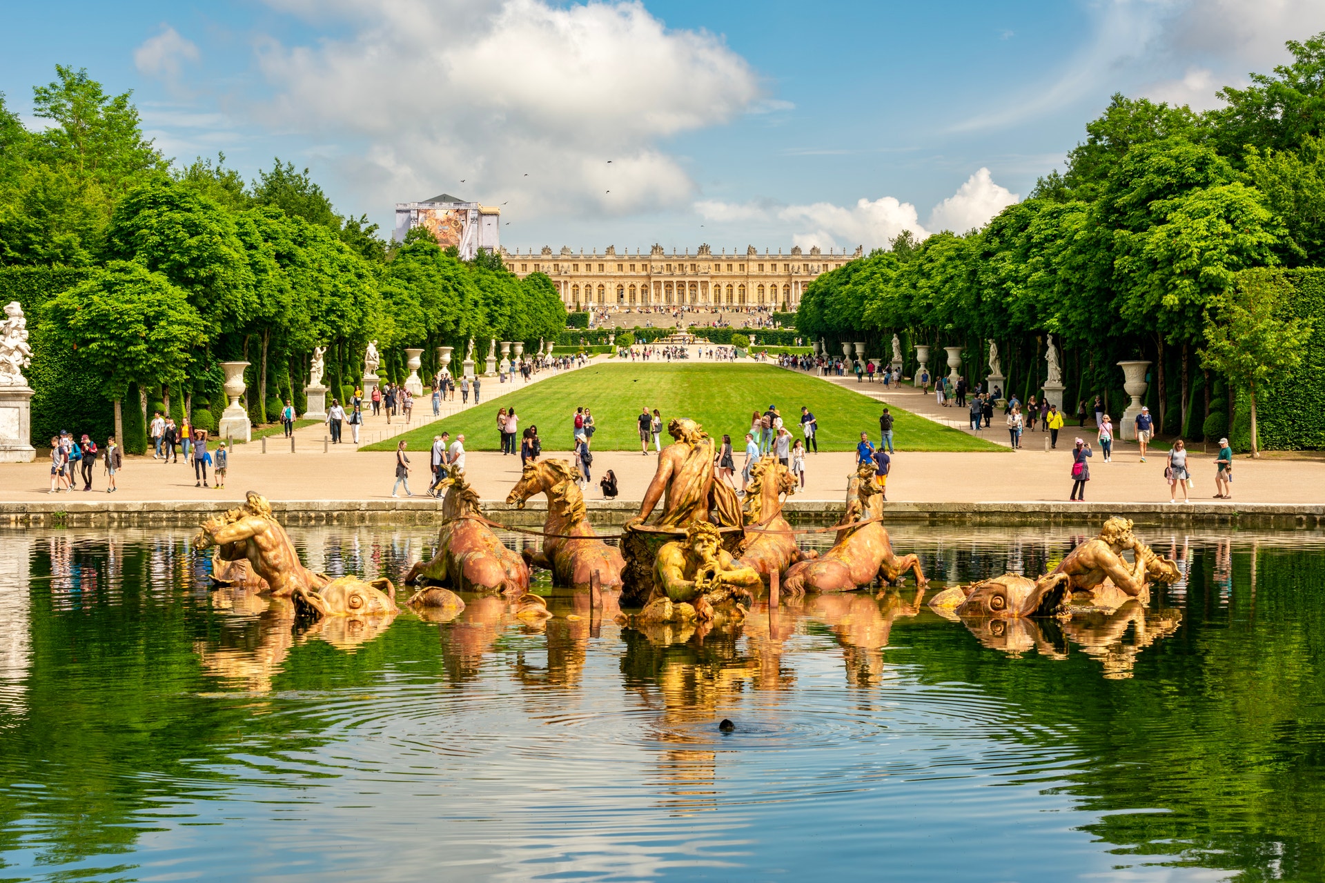 Slottet i Versailles med trädgårdar, en fontän med guldstatyer och besökare som promenerar på gräsmattan.