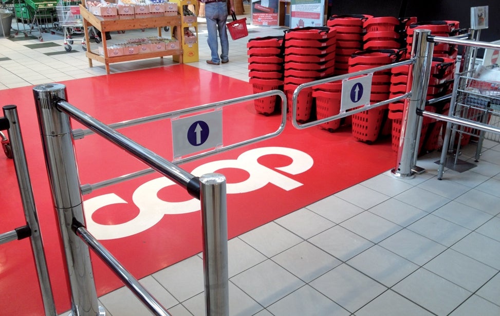 En butiksmiljö med röda köpvagnar och väggmarkering med co-op logotyp. Kövägen är markerad med röda mattor och metallbarriärer med nummertavlor för köhantering.