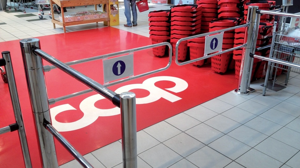 En butiksmiljö med röda köpvagnar och väggmarkering med co-op logotyp. Kövägen är markerad med röda mattor och metallbarriärer med nummertavlor för köhantering.