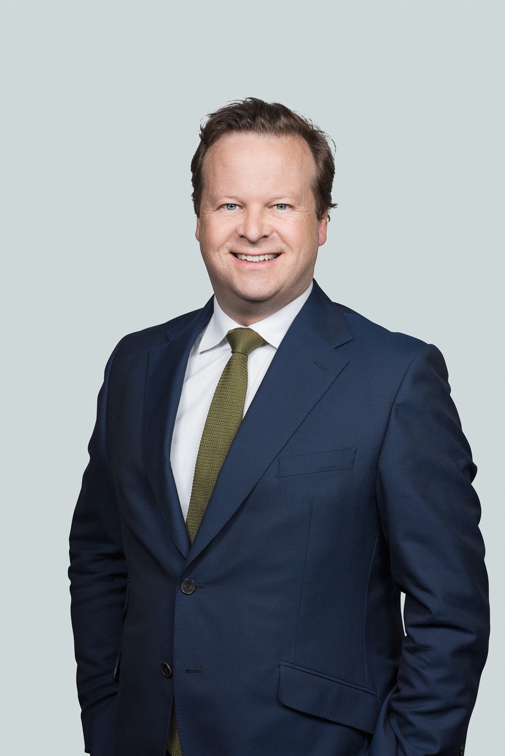 Porträttfoto av en man i marinblå kostym och grön slips mot ljusgrå bakgrund.