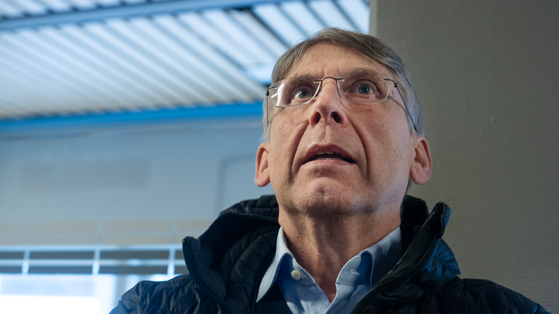 Porträtt av en man med glasögon och mörk jacka som tittar uppåt i en modern inomhusmiljö.