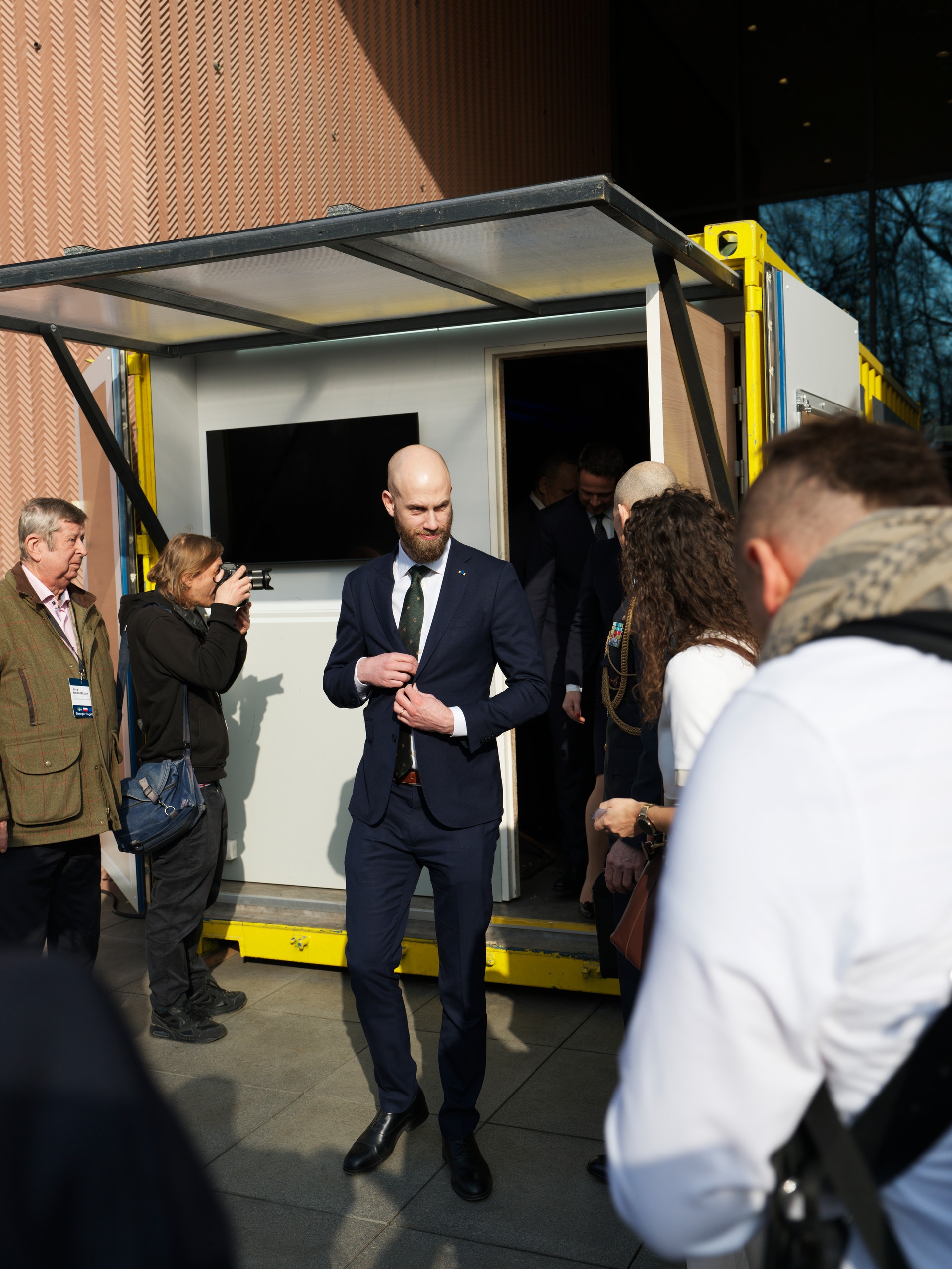 Man i mörk kostym står framför en vit containerbyggnad omgiven av människor vid ett officiellt evenemang.