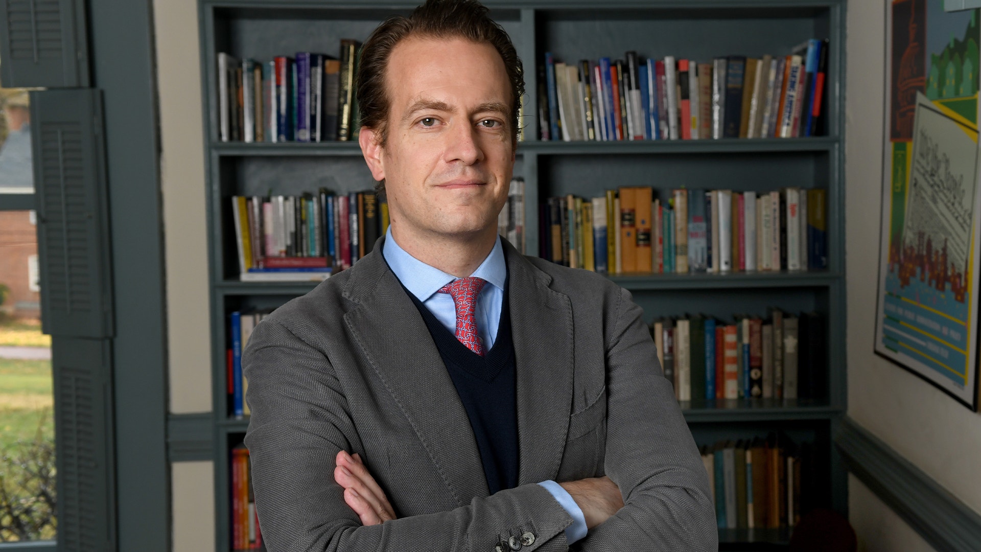 Man i grå kavaj och blå skjorta står framför en bokhylla med armarna i kors.