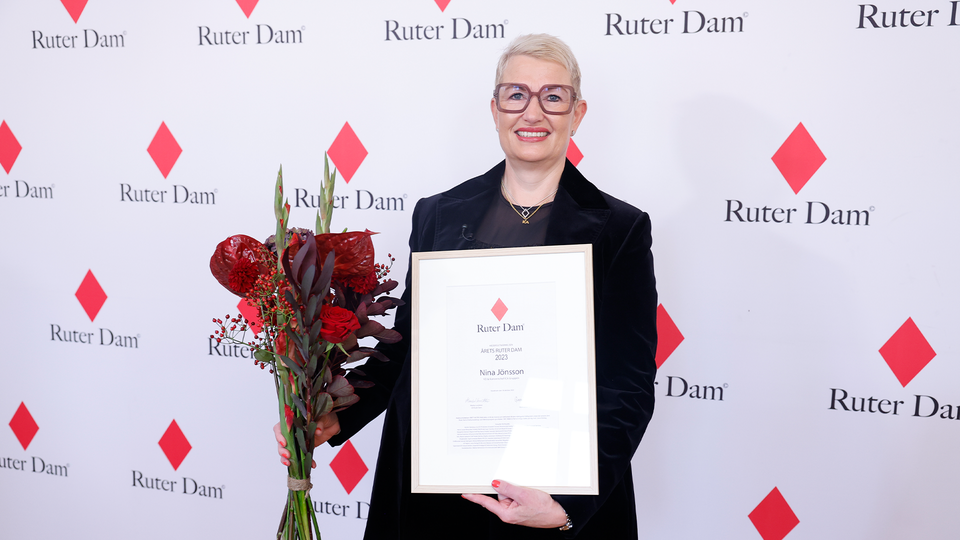 Nina Jönsson, en blond korthårig kvinna med stora mörka glasögon och rött läppstift, står framför en vägg där det står "Ruter dam". Hon håller ett diplom och en blombukett.