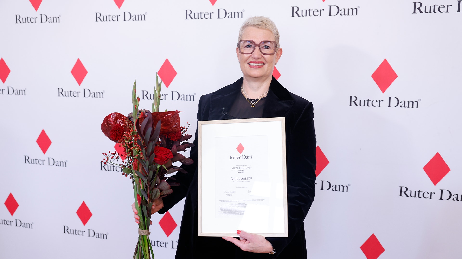 Nina Jönsson, en blond korthårig kvinna med stora mörka glasögon och rött läppstift, står framför en vägg där det står "Ruter dam". Hon håller ett diplom och en blombukett.
