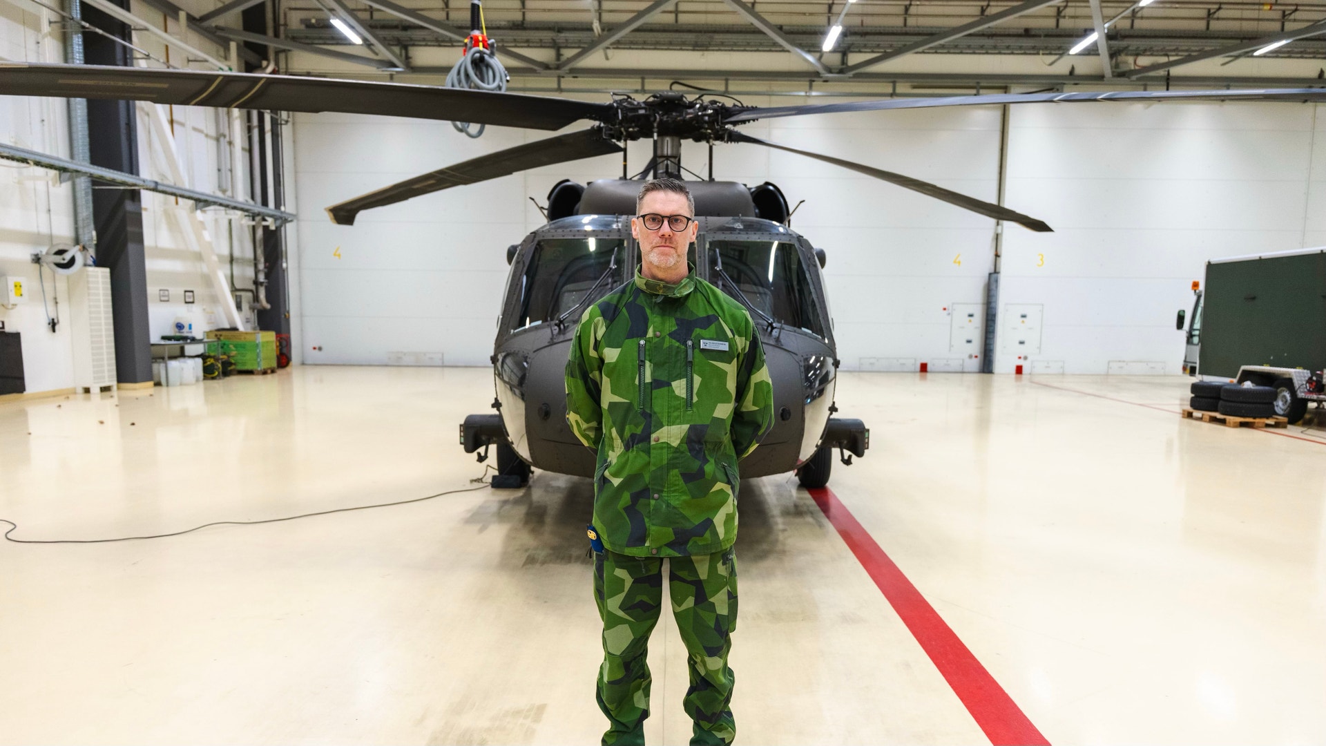 Militär pilot i kamouflageuniform står framför en helikopter i en hangar.