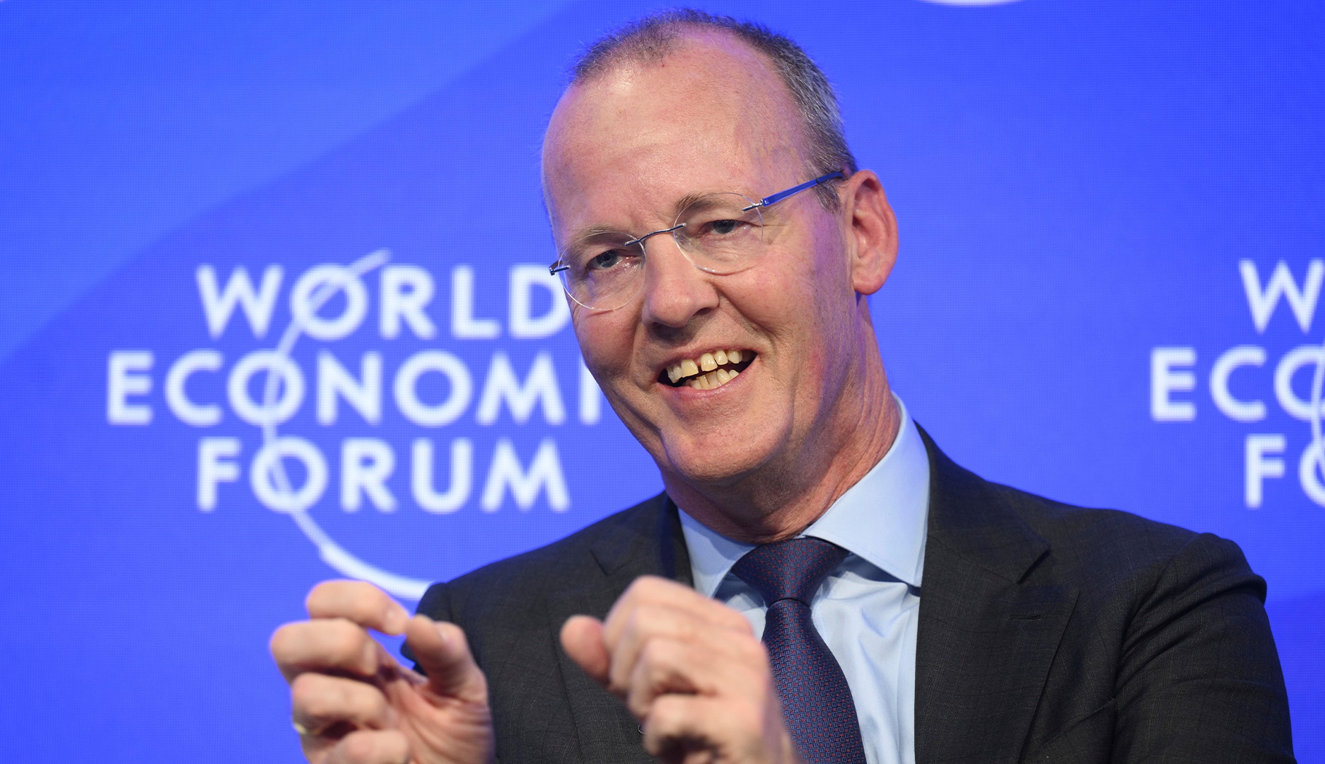 Leende man i glasögon och kostym som gestikulerar med händerna framför en blå bakgrund med World Economic Forums logotyp.