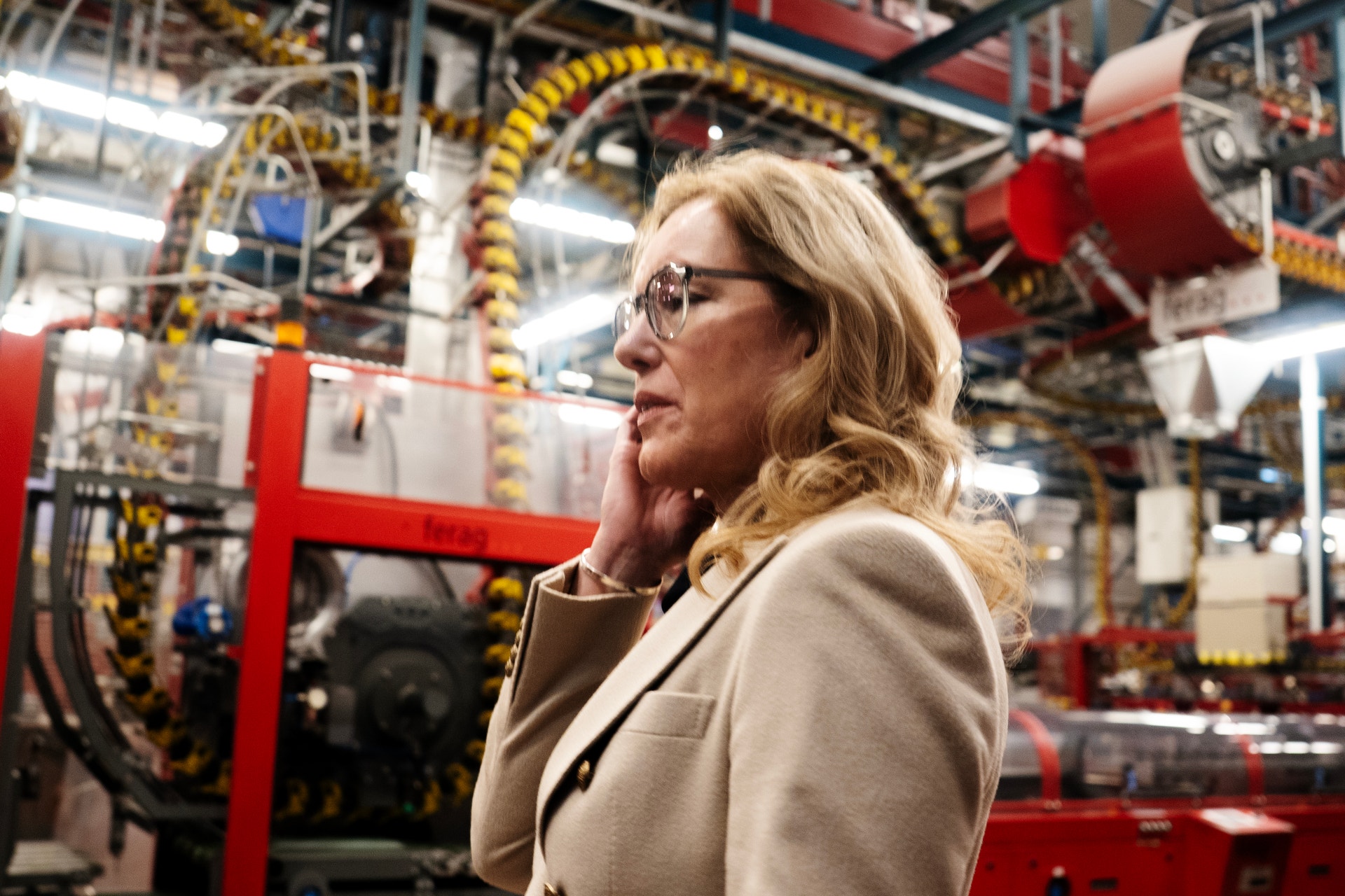 Kvinna i glasögon och beige blazer står i en industriell fabrik omgiven av maskiner och utrustning.