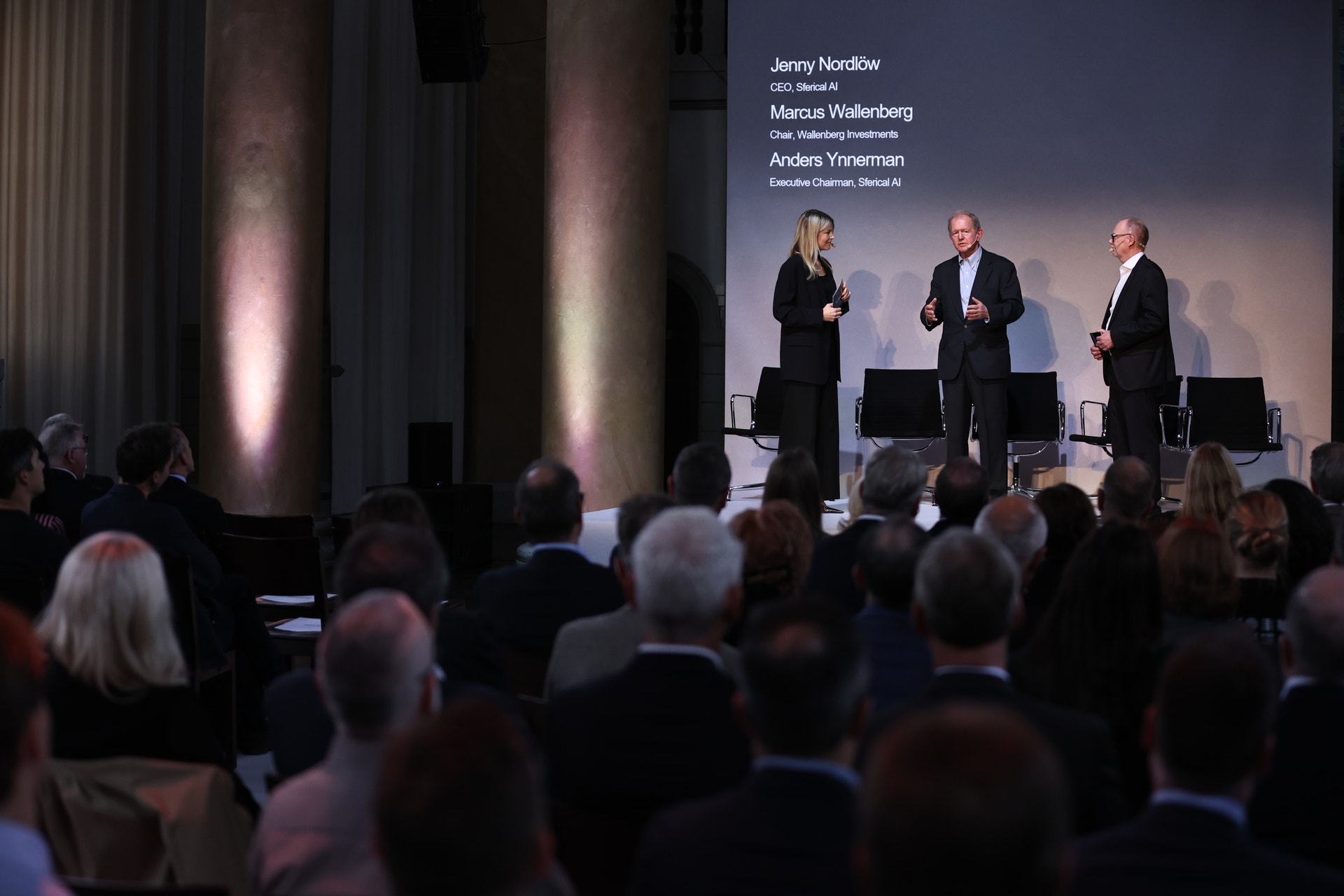 Tre personer i mörka kläder står på en scen och pratar framför en publik. Bakom dem visas en presentationsskärm.