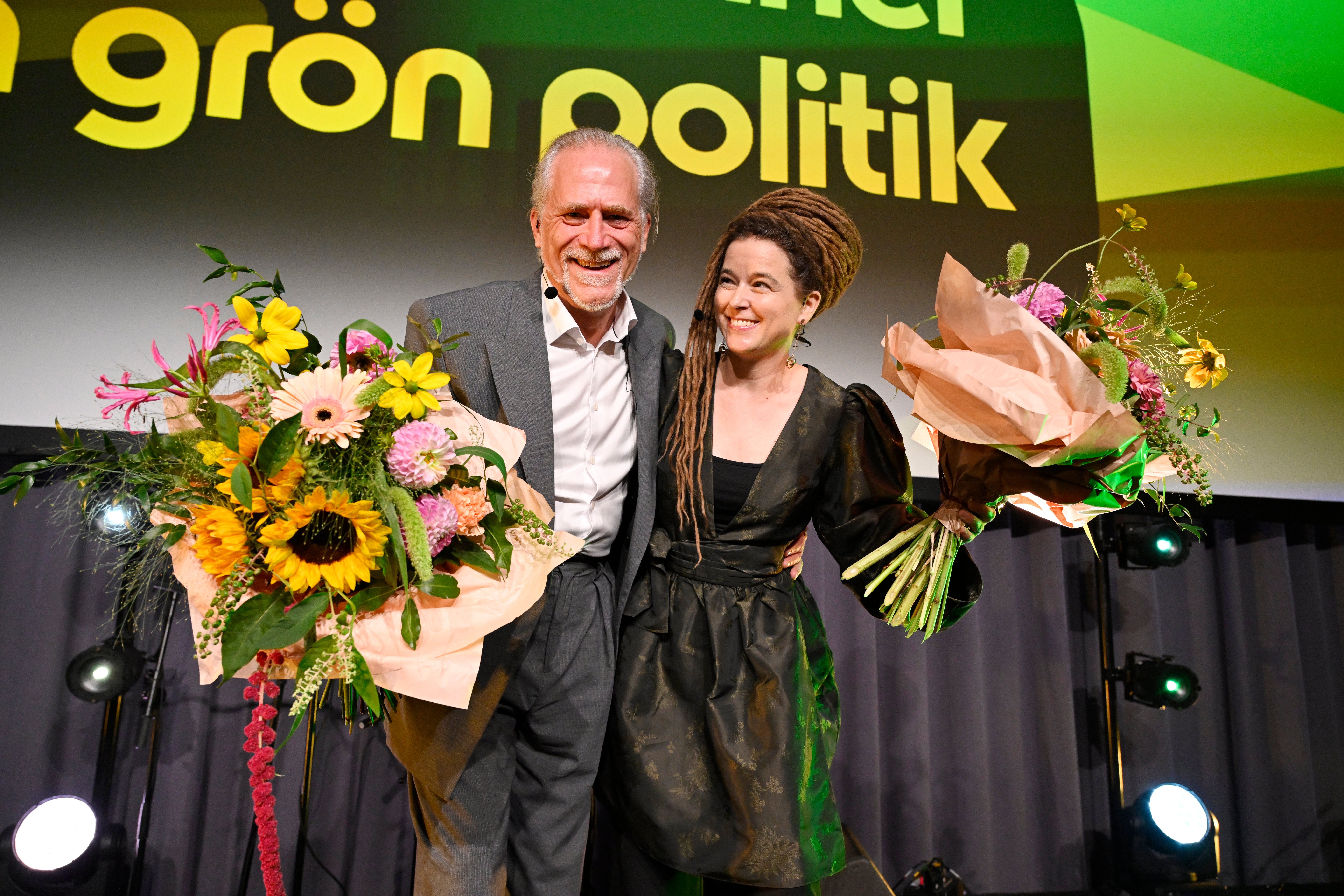 En man och en kvinna står på en scen och håller varsin blombukett, med en grön bakgrundsskylt som nämner politik.
