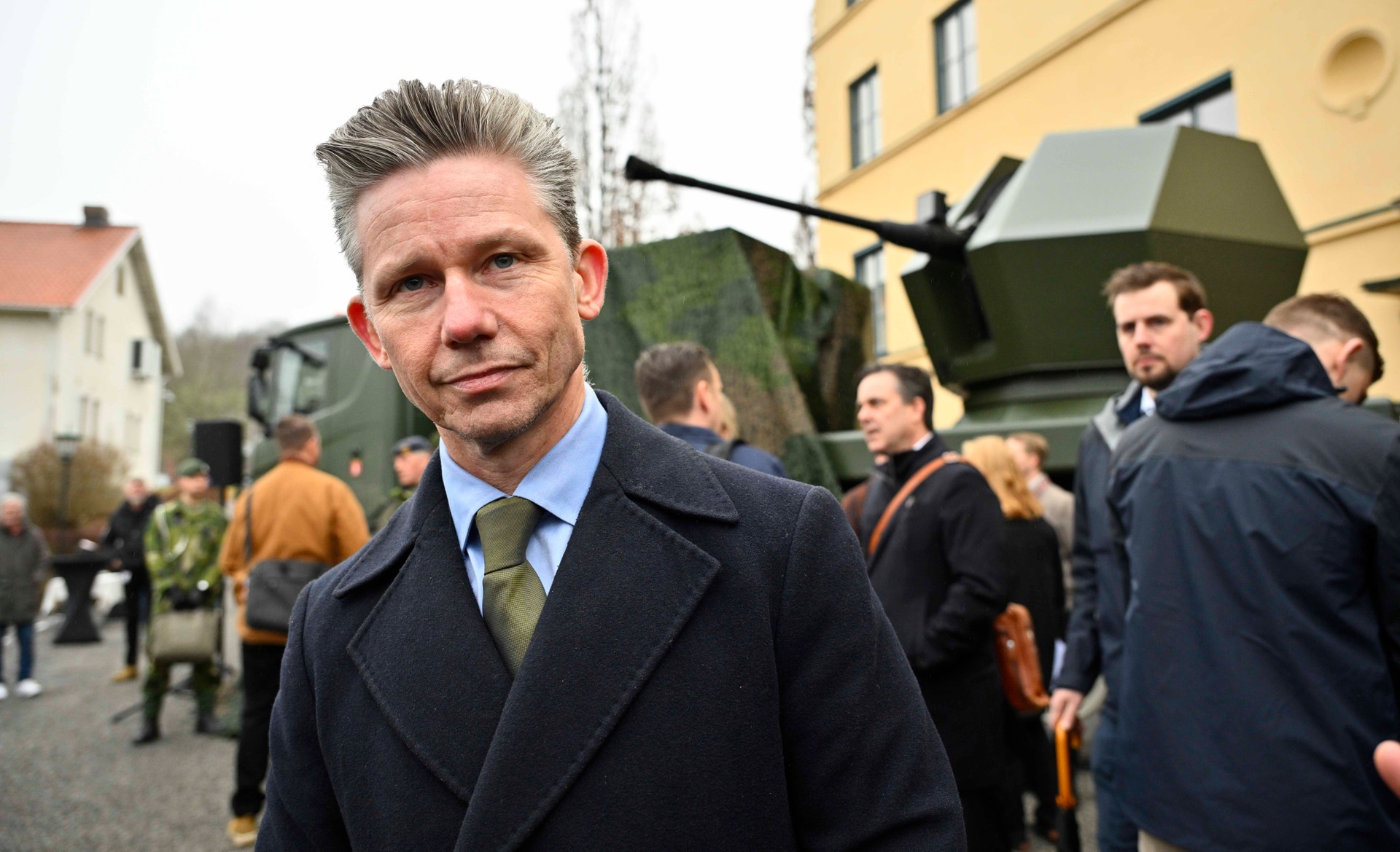Porträtt av en man i mörk kostym som står framför militär utrustning och andra personer vid ett event.