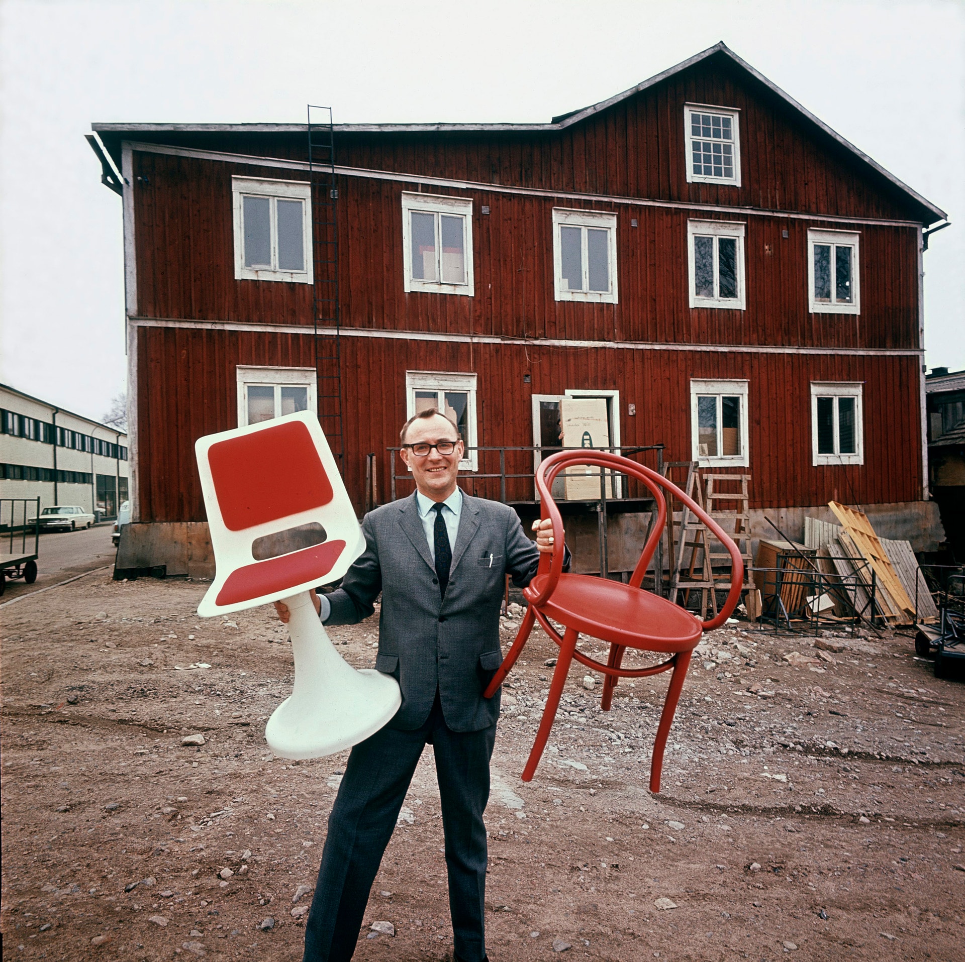 Man i kostym poserar med moderna möbler framför rött trähus från 1970.
