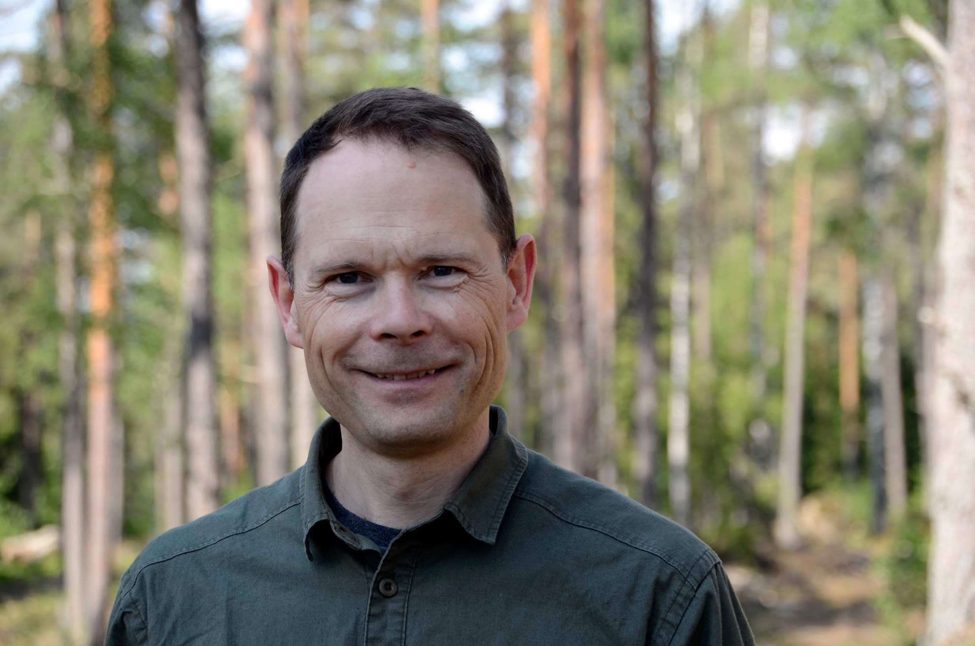 Porträtt av en man i grön skjorta som ler, med skog i bakgrunden.