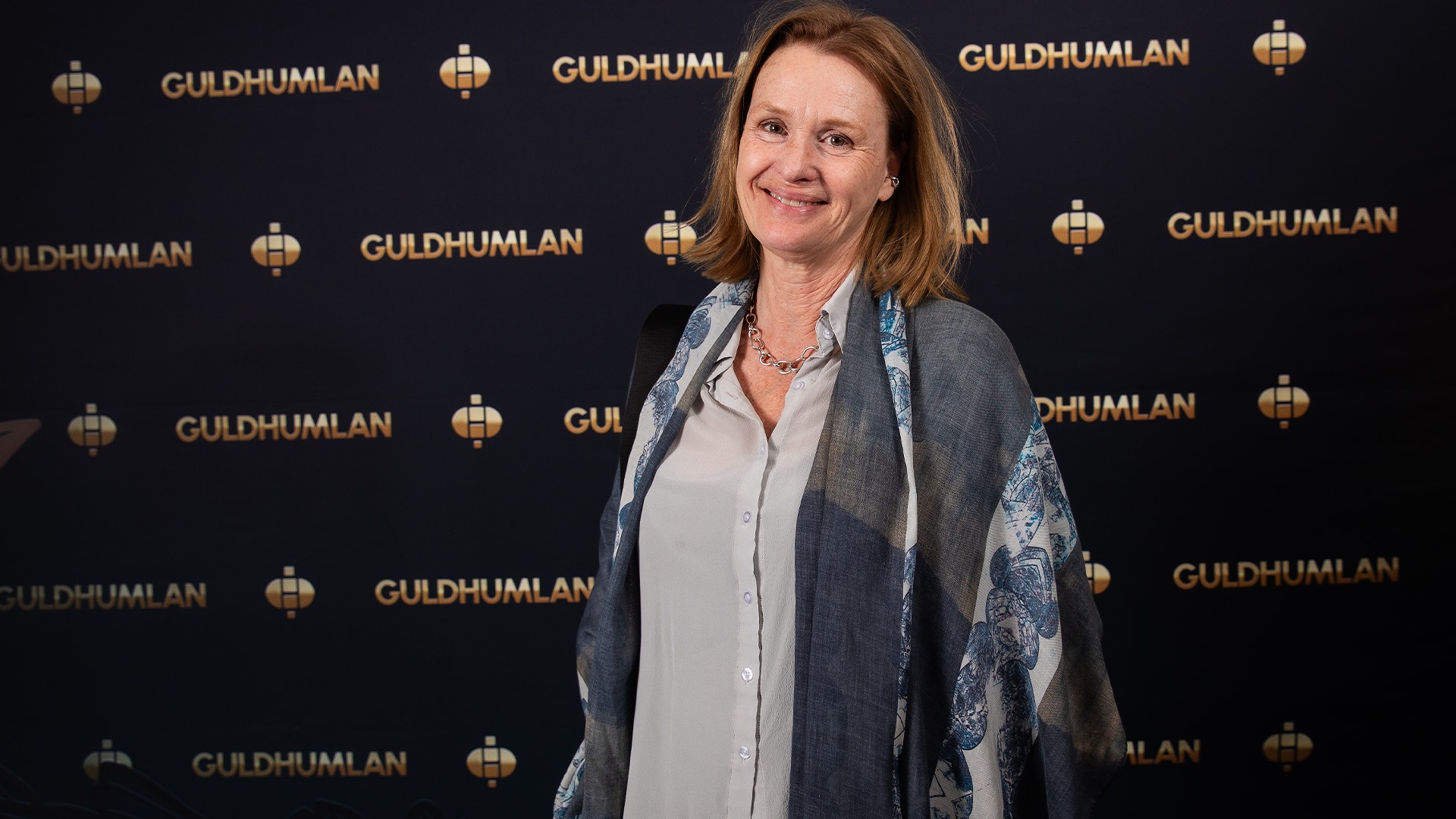 En kvinna står framför en mörk vägg med Guldhumlan-logotyper. Hon bär en ljusblå skjorta och en grå cardigan med blå detaljer.