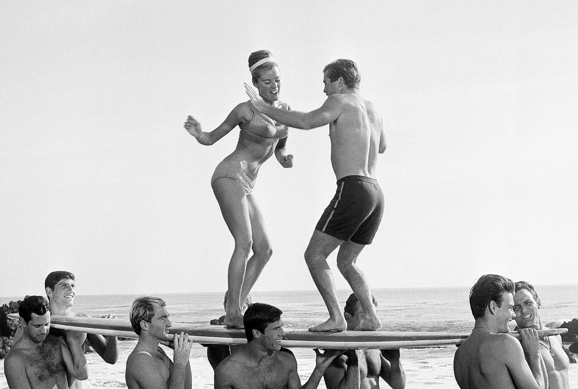 Svartvit fotografi från en strand. Två personer dansar på en träbräda som hålls upp av flera andra personer. Havet och horisonten syns i bakgrunden.