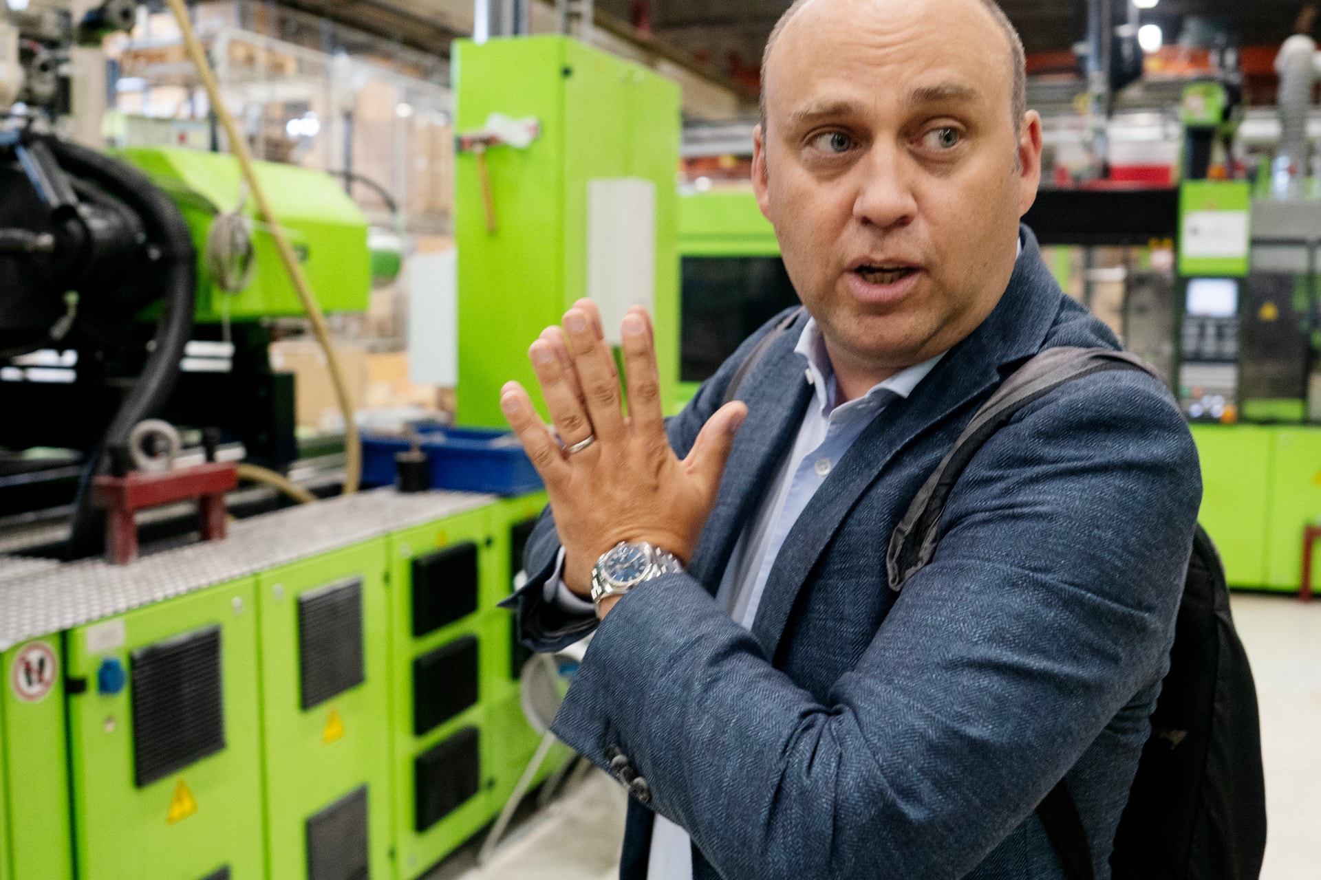 En man i blå skjorta och jacka gestikulerar medan han pratar i en fabrik omgiven av grön industriell utrustning.