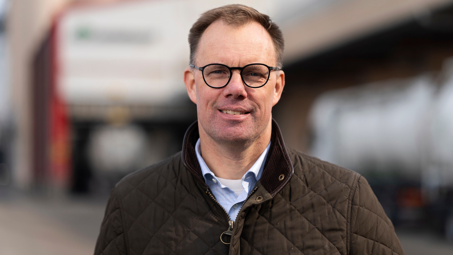 Porträtt av en man med glasögon och brun jacka framför en industribyggnad.