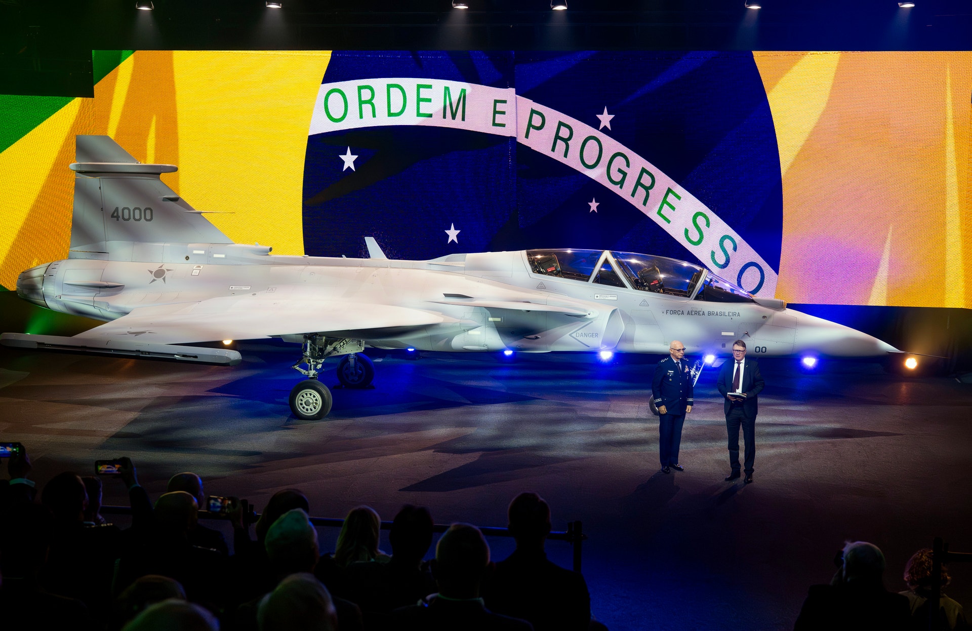 Ett modernt stridsflygplan presenteras på en ceremoni framför den brasilianska flaggan. Flygplanet är belyst med blå ljus och människor tittar på från förgrunden.