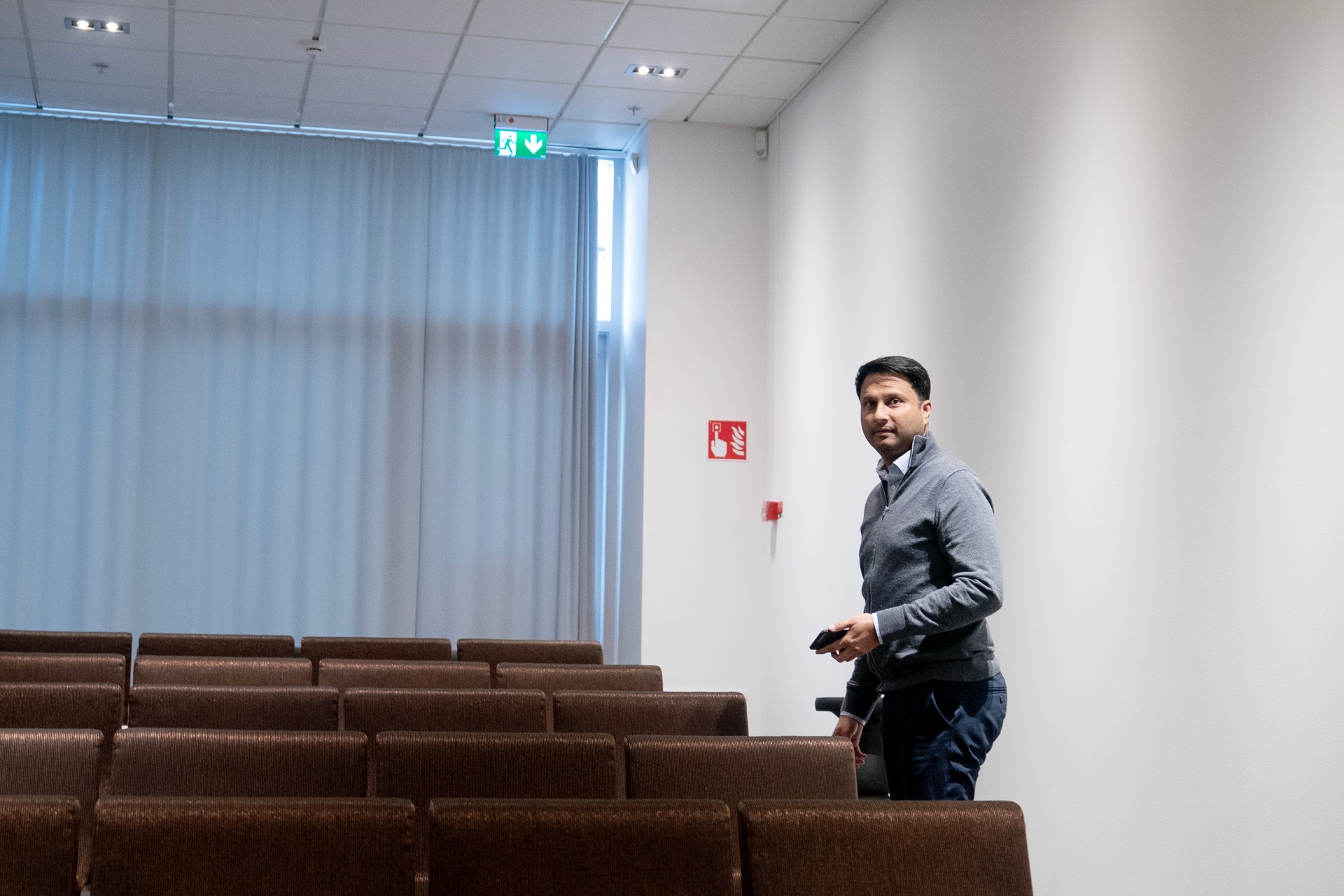 Porträtt av Rishit Jhunjhunwala, VD för Truecaller, som står i en auditorium i Stockholm. Han är klädd i en mörkblå skjorta och står framför bruna säten. I bakgrunden syns stora fönster med vertikala gardiner, vita väggar och en nödutgångsskylt.