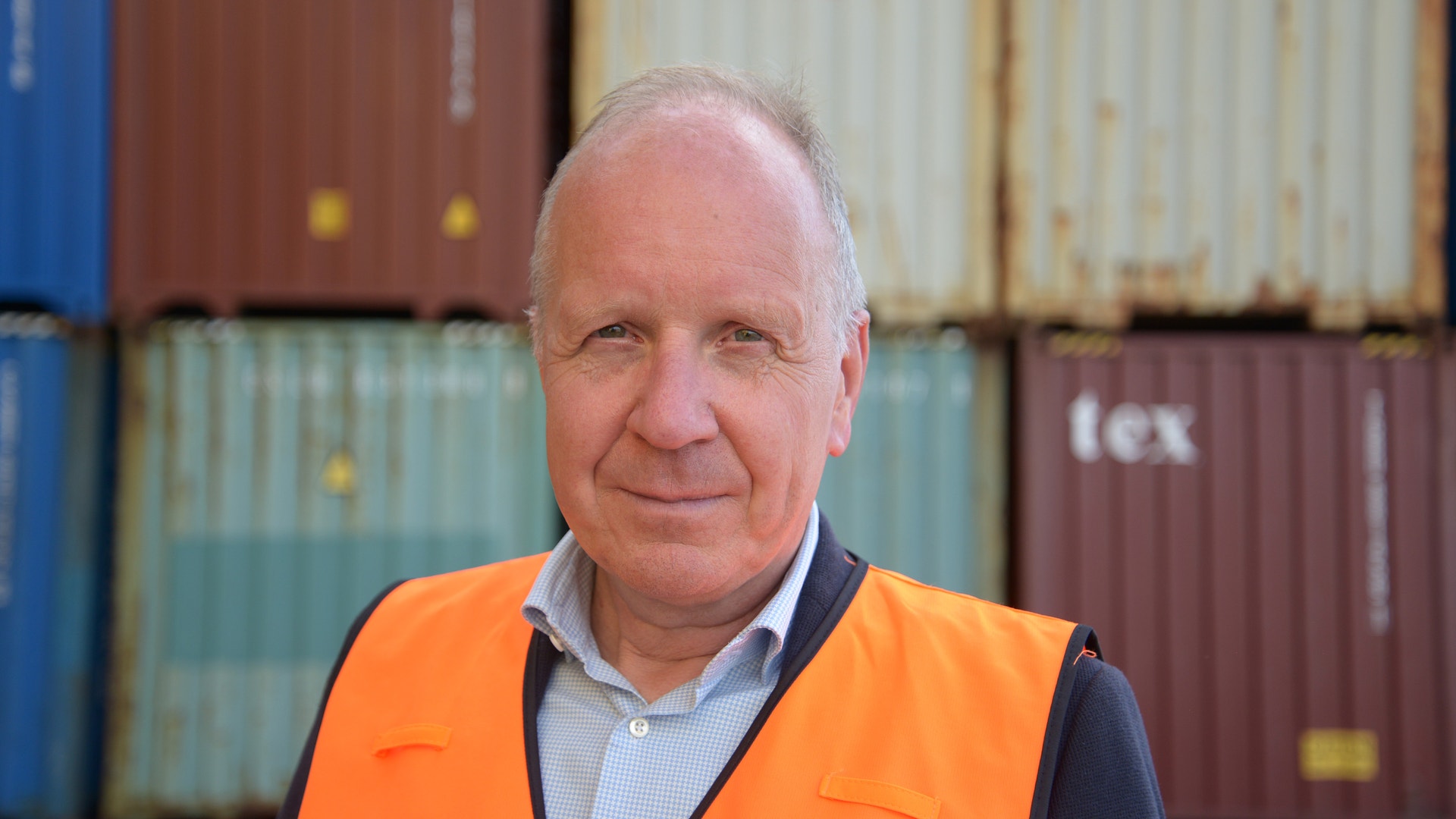Porträtt av en man i orange säkerhetsvest som står framför lastcontainrar.