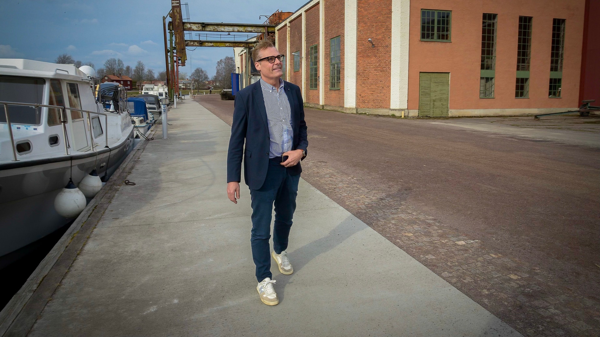 En man i mörk kavaj går på en gata framför en röd tegelbyggnad. Båtar syns i bakgrunden.