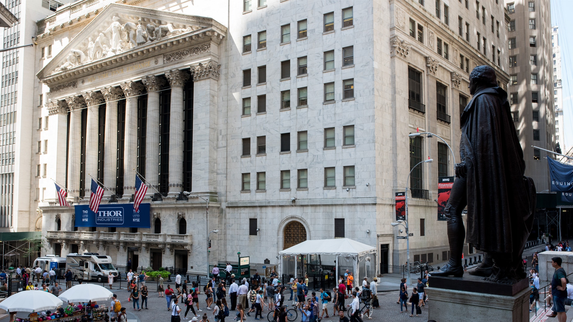 New York Stock Exchange-byggnaden på Wall Street med vita kolonner, amerikanska flaggor och en staty i förgrunden. Många människor går på platsen framför byggnaden.