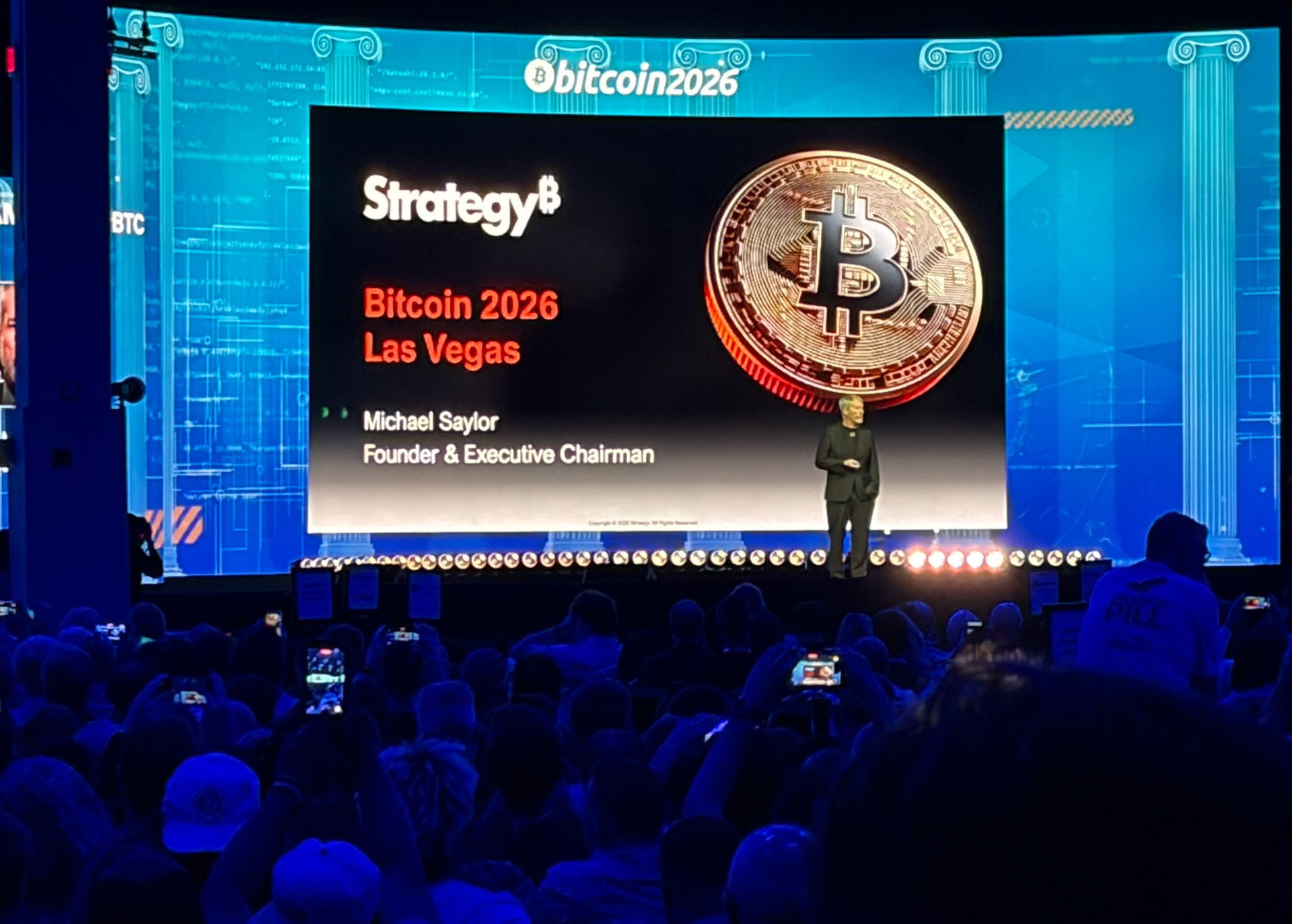 Konferenspresentation på Bitcoin 2026 i Las Vegas. En talare står på scenen framför en stor skärm som visar en gyllene Bitcoin-mynt och presentationen från Strategy8 med Michael Saylor, Founder & Executive Chairman. Publiken filmar med sina telefoner.