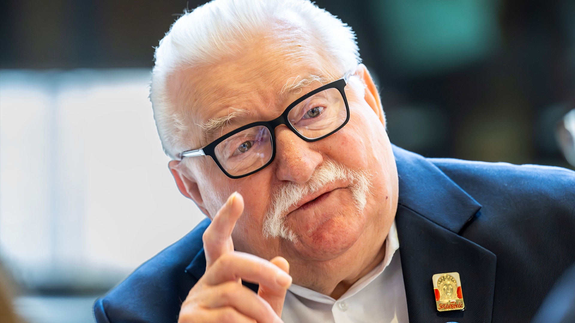 Äldre man med vitt hår och glasögon i mörkblå kostym som gestikulerar medan han talar.