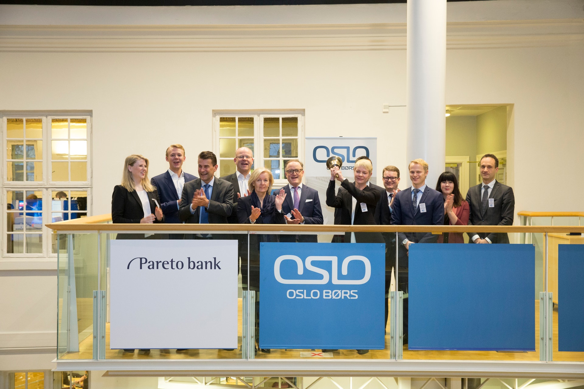 Gruppfoto av omkring 15 personer i formell klädsel som står på en balkong med banderoller för Pareto bank och Oslo Børs.