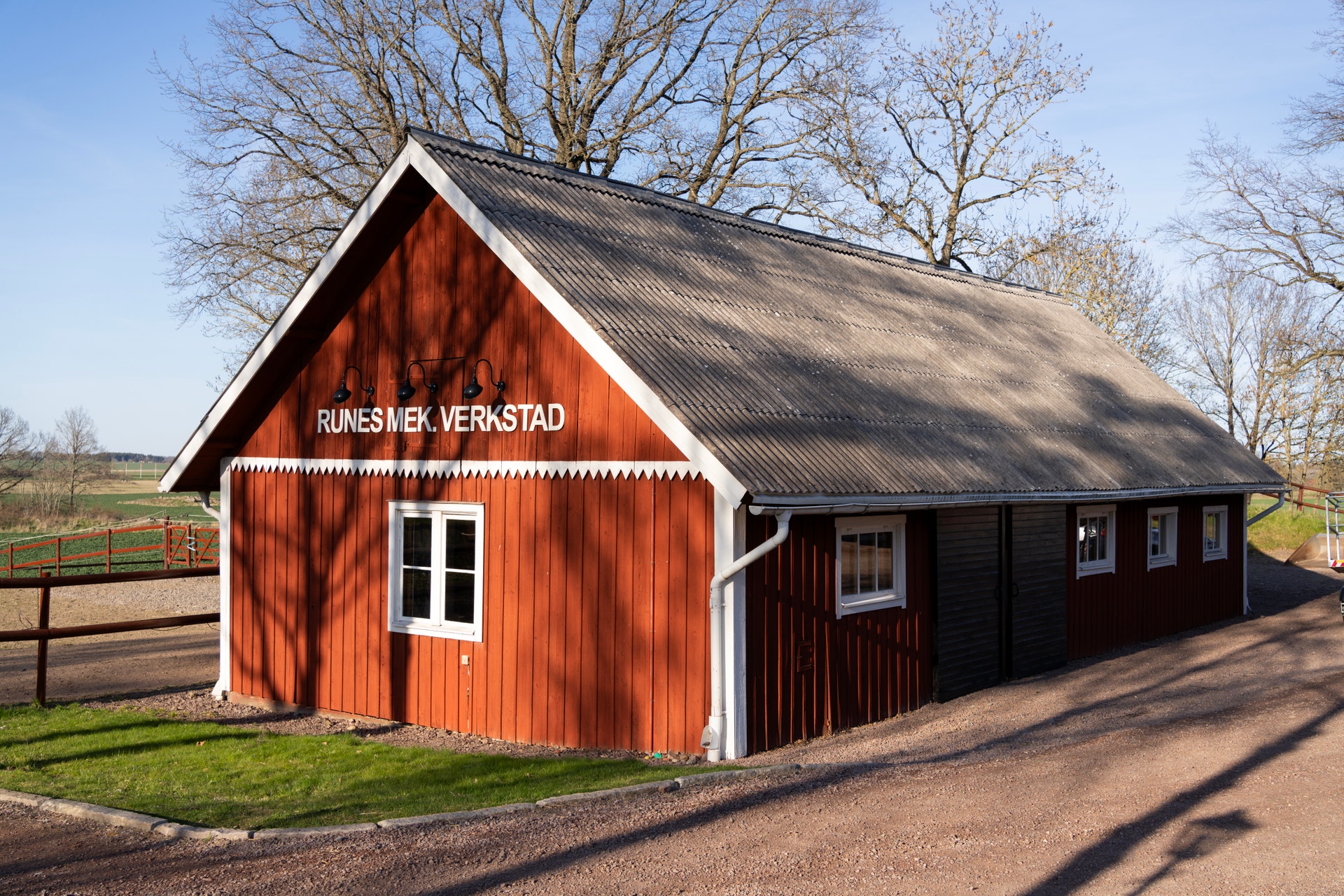 Röd trähus med vita detaljer och naturligt taktäcke. Huset är märkt 'Runes Mek. Verkstad' och ligger i en landsbygdsmiljö omgiven av träd och gräs.