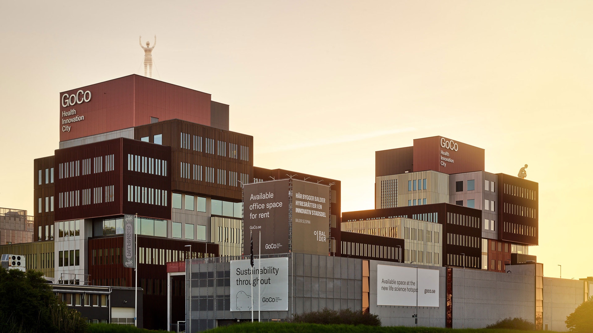 Moderna kontorsbyggnader i rödbrunt och grått med texten GoCo Health Innovation City, fotograferade i varmt kvällsljus.