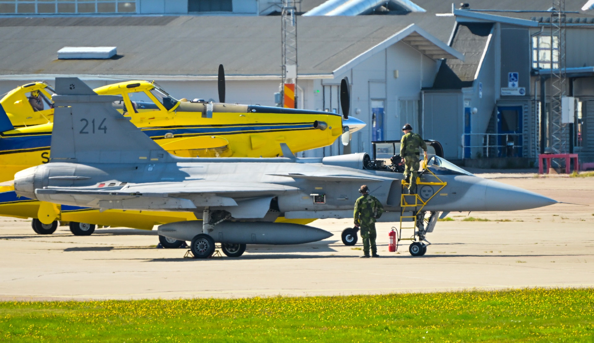 Ett stridsflygplan av typen JAS 39 Gripen står parkerat på en flygplats med militär personal i närheten, samt gula propellerplan i bakgrunden.