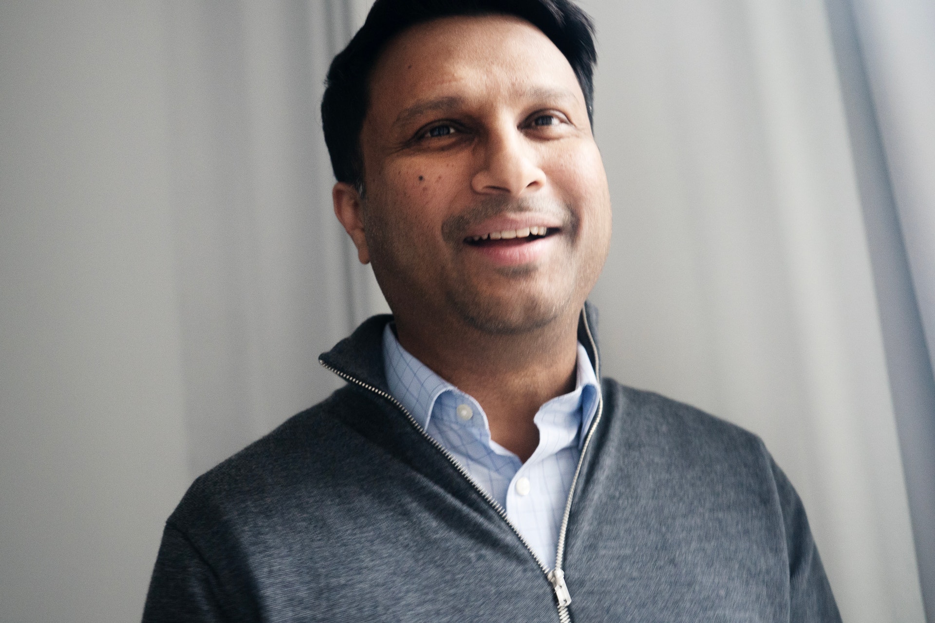 Porträttfoto av en man med mörkt hår som ler mot kameran. Han bär en grå cardigan över en ljusblå skjorta och är fotograferad mot en vit bakgrund.