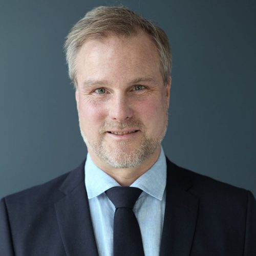 Porträtt av en man i mörk kostym och ljusblå skjorta med mörkblå slips mot neutral grå bakgrund.