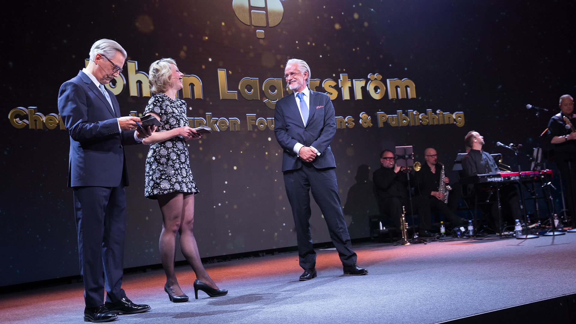 Tre personer i formell klädsel på en belyst scen under en gala. I bakgrunden syns en orkester och text med Johan Lagerströms namn.