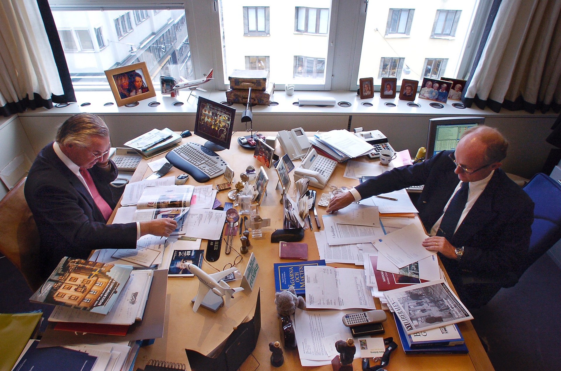 Två män i kostym arbetar vid ett skrivbord fullt av papper och dokument i ett kontorsrum med stora fönster mot en stadsutsikt.