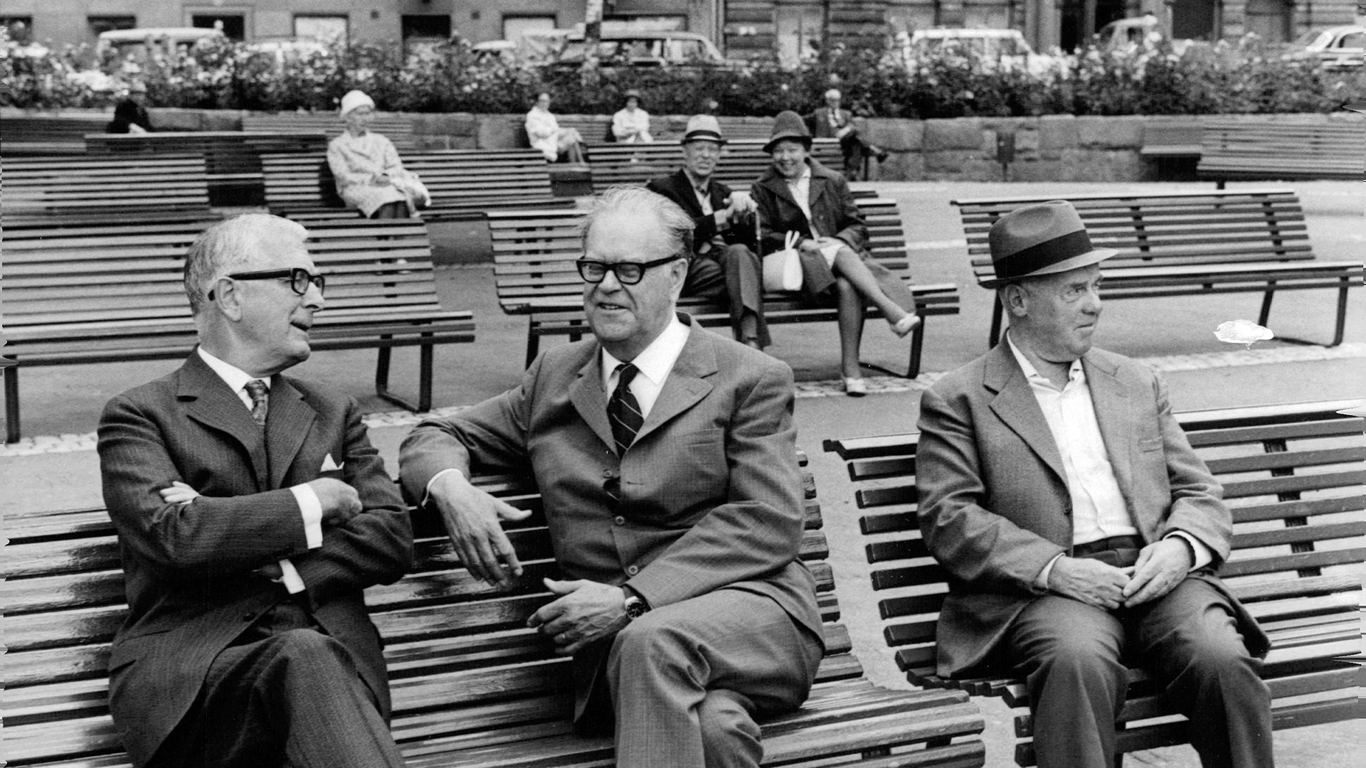 Tre män i kostym sitter på en parkbänk. Svartvit fotografi från mitten av 1900-talet.