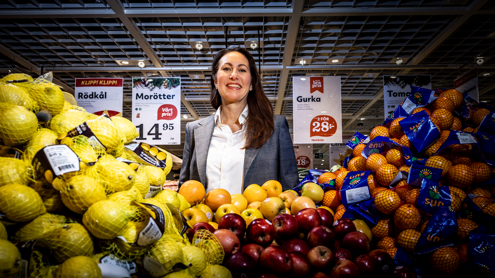 En kvinna i grå kavaj och vit skjorta står i en livsmedelsbutiks fruktavdelning omgiven av gula äpplen, röda äpplen och apelsiner. Prisskyltarna och svenska skyltar är synliga i bakgrunden, tillsammans med metalliska takbalkar som är typiska för en supermarknad.
