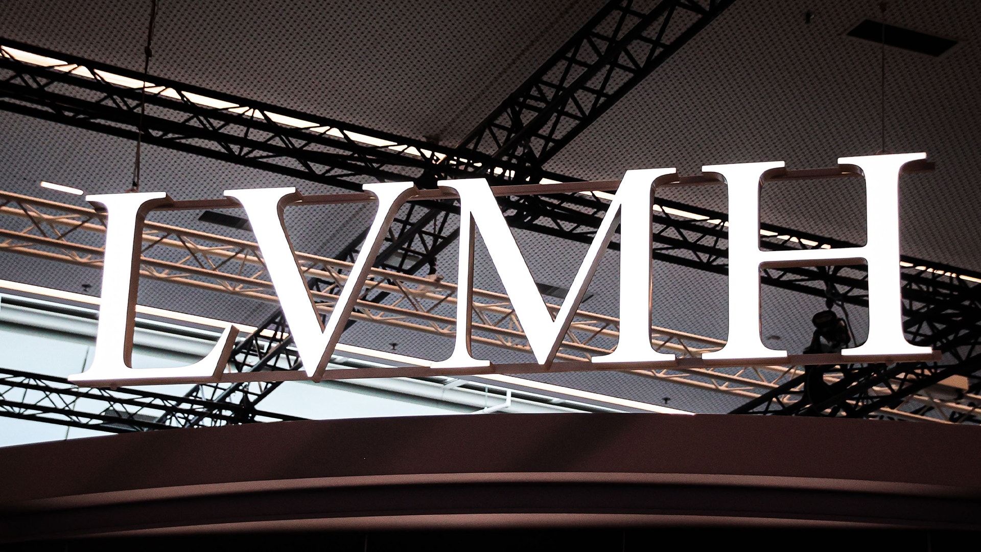 Vit skylt med texten LVMH uppsatt under ett mörkt tak med metallbjälkar.