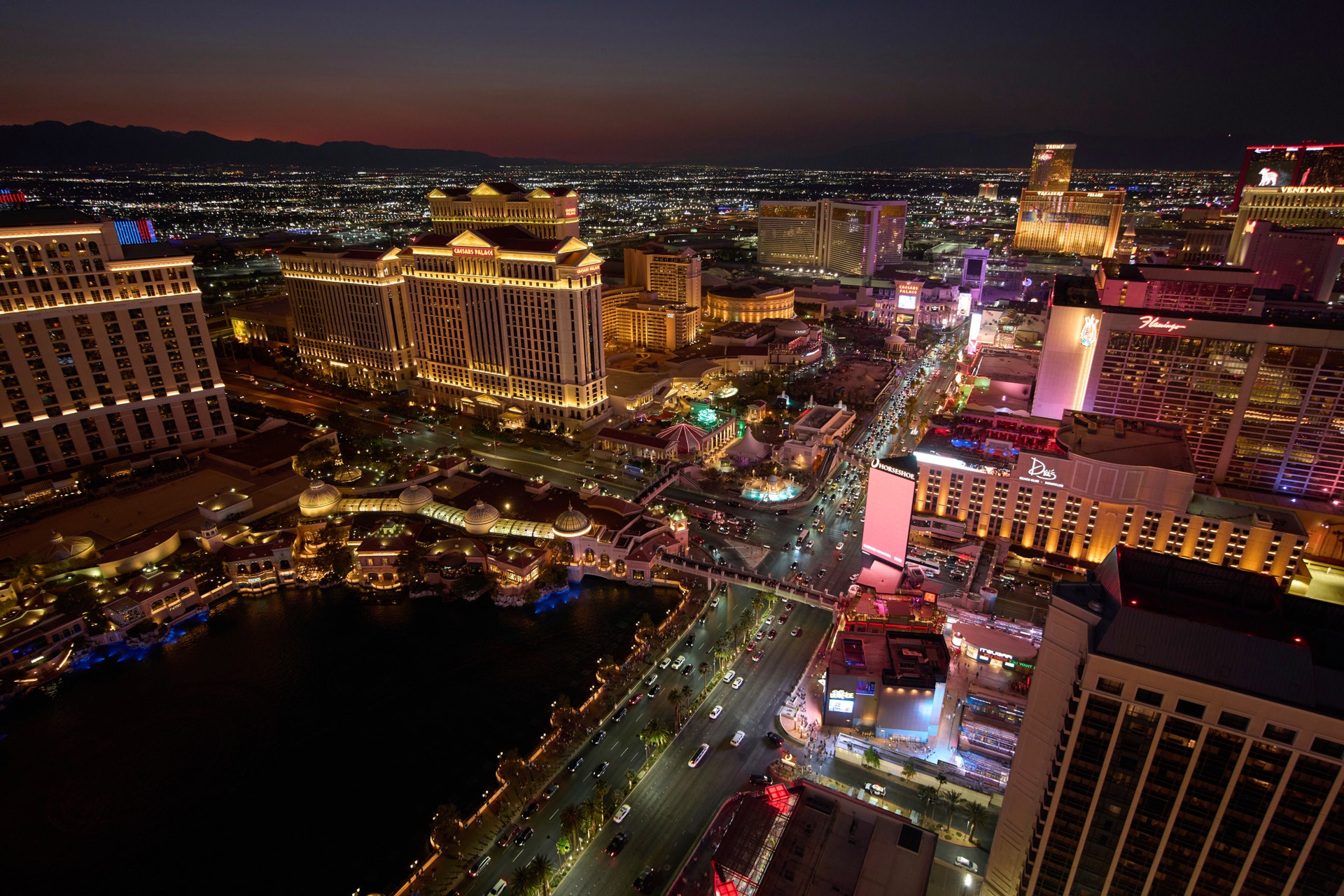 Nattvy över Las Vegas Strip med upplysta hotell och kasinon. Stora byggnader i gult och rött ljus omger en mörkblå sjö, med bergen synliga i bakgrunden under en skymningshimmel.
