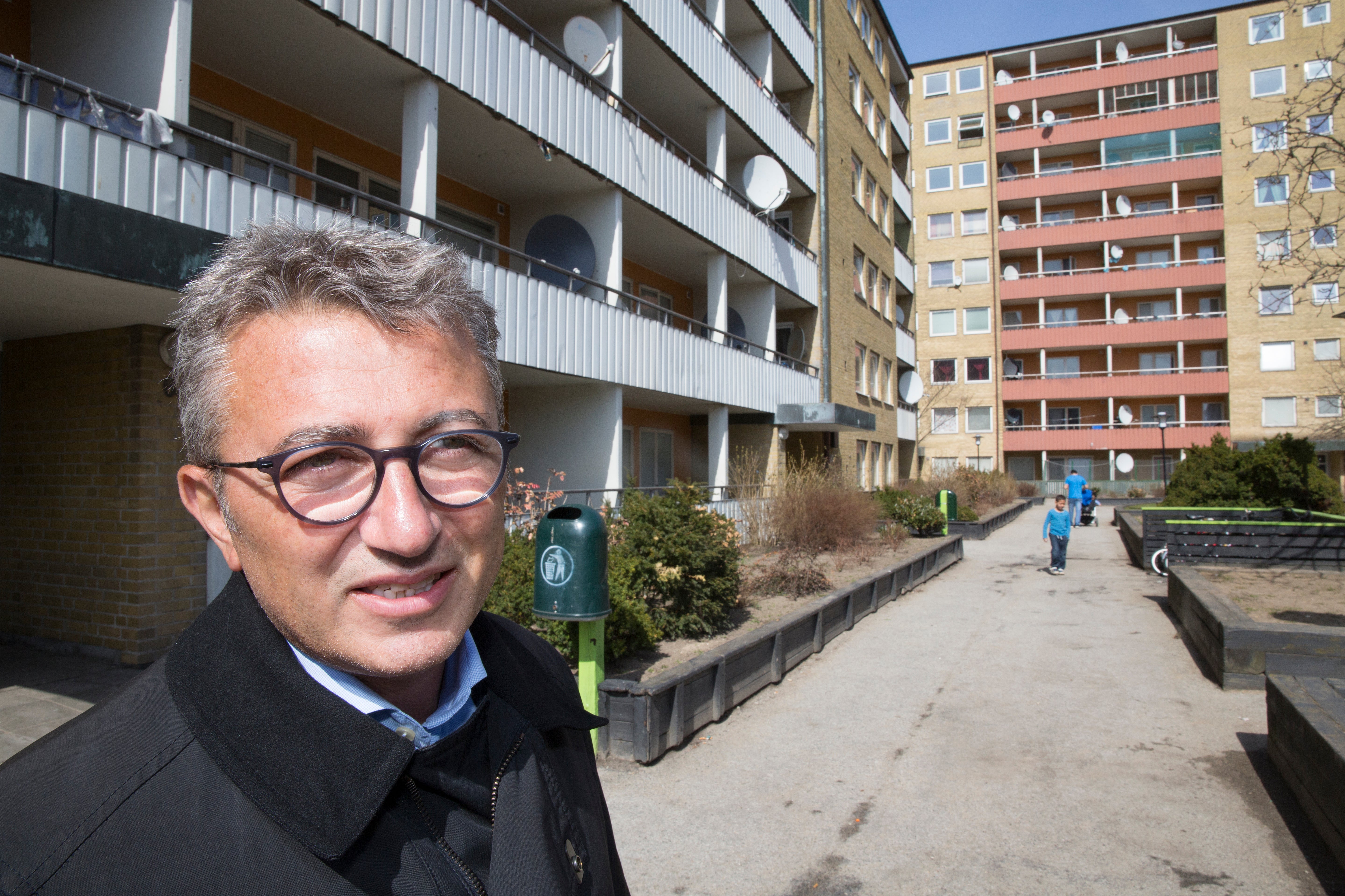 En man med grått hår och glasögon, klädd i svart jacka, står utomhus framför höga bostadshus av tegel och betong.