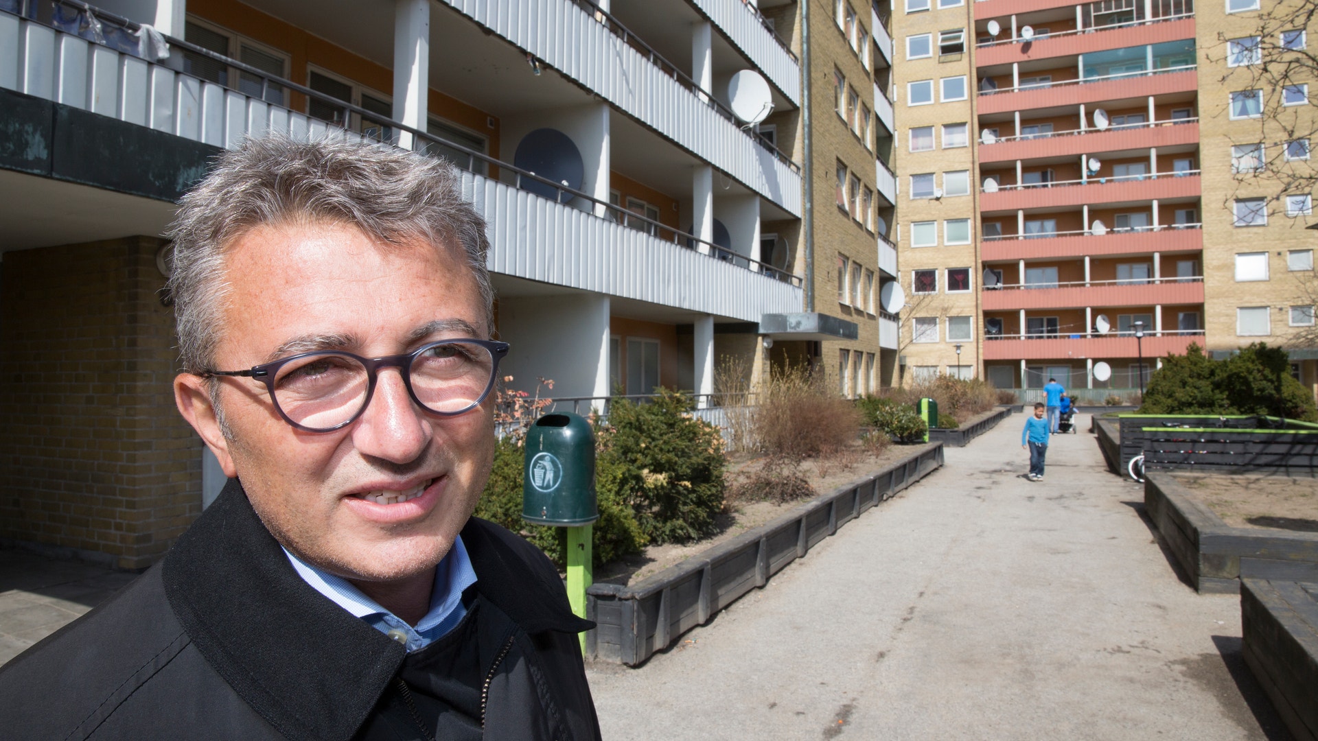 En man med grått hår och glasögon, klädd i svart jacka, står utomhus framför höga bostadshus av tegel och betong.